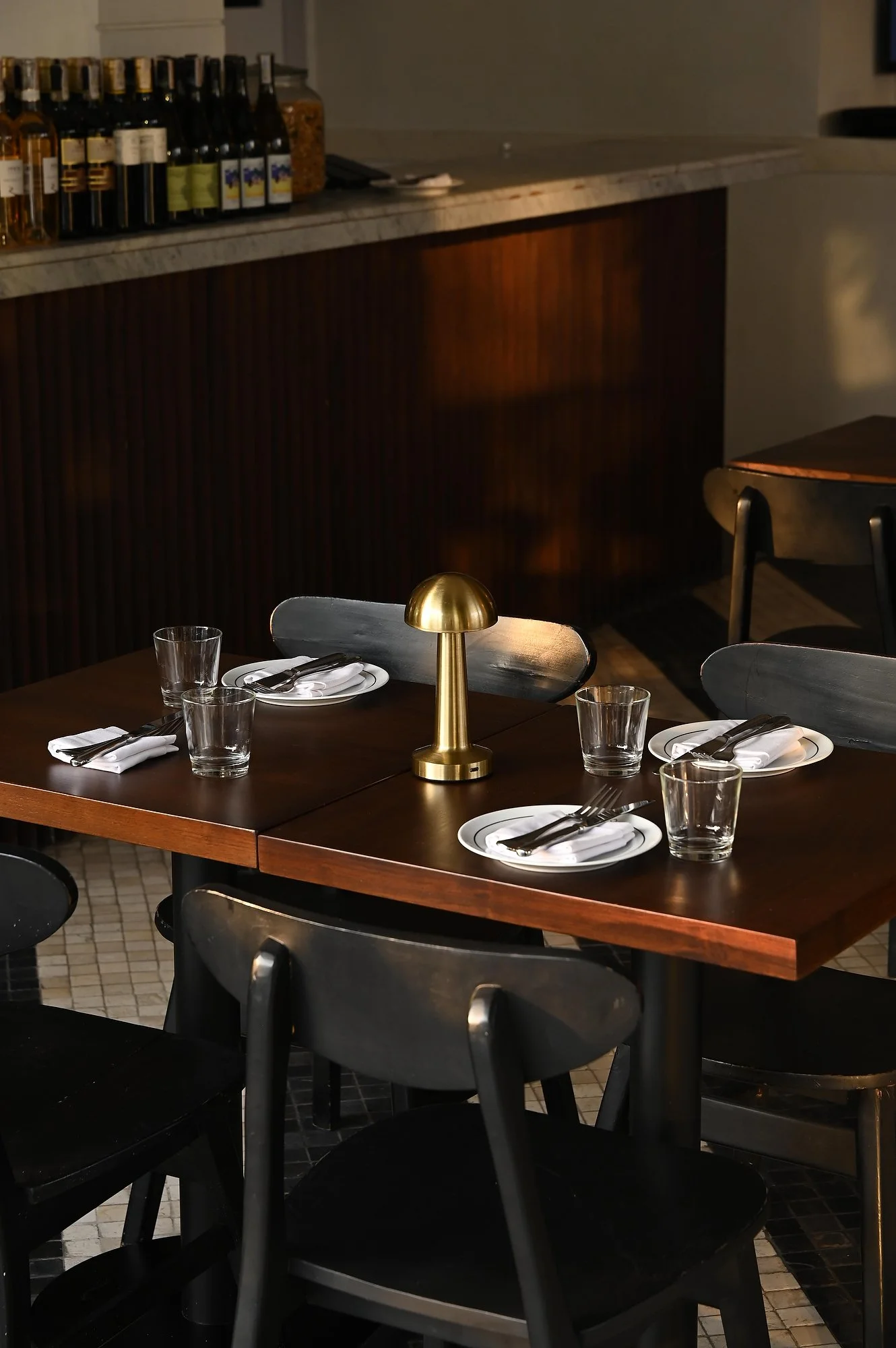 Table set for dining with white plates, silverware, napkins, empty glasses, a gold lamp, and dark chairs in a dimly lit restaurant.