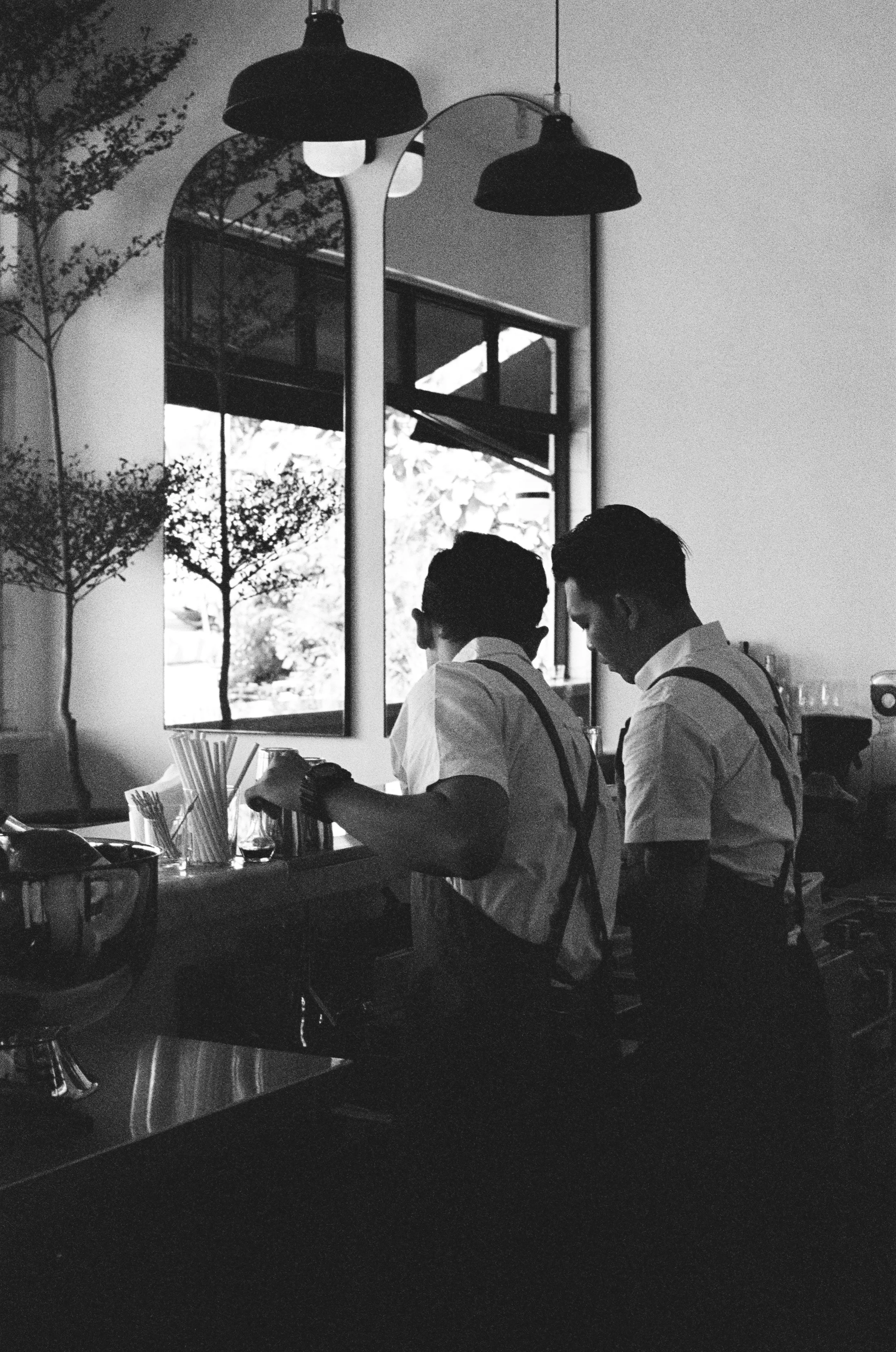 Two men wearing white shirts and aprons, likely chefs, preparing food at a kitchen counter with various utensils, in a restaurant or cafe. They are working in front of a large mirror, with a window showing trees outside, illuminated by hanging pendan