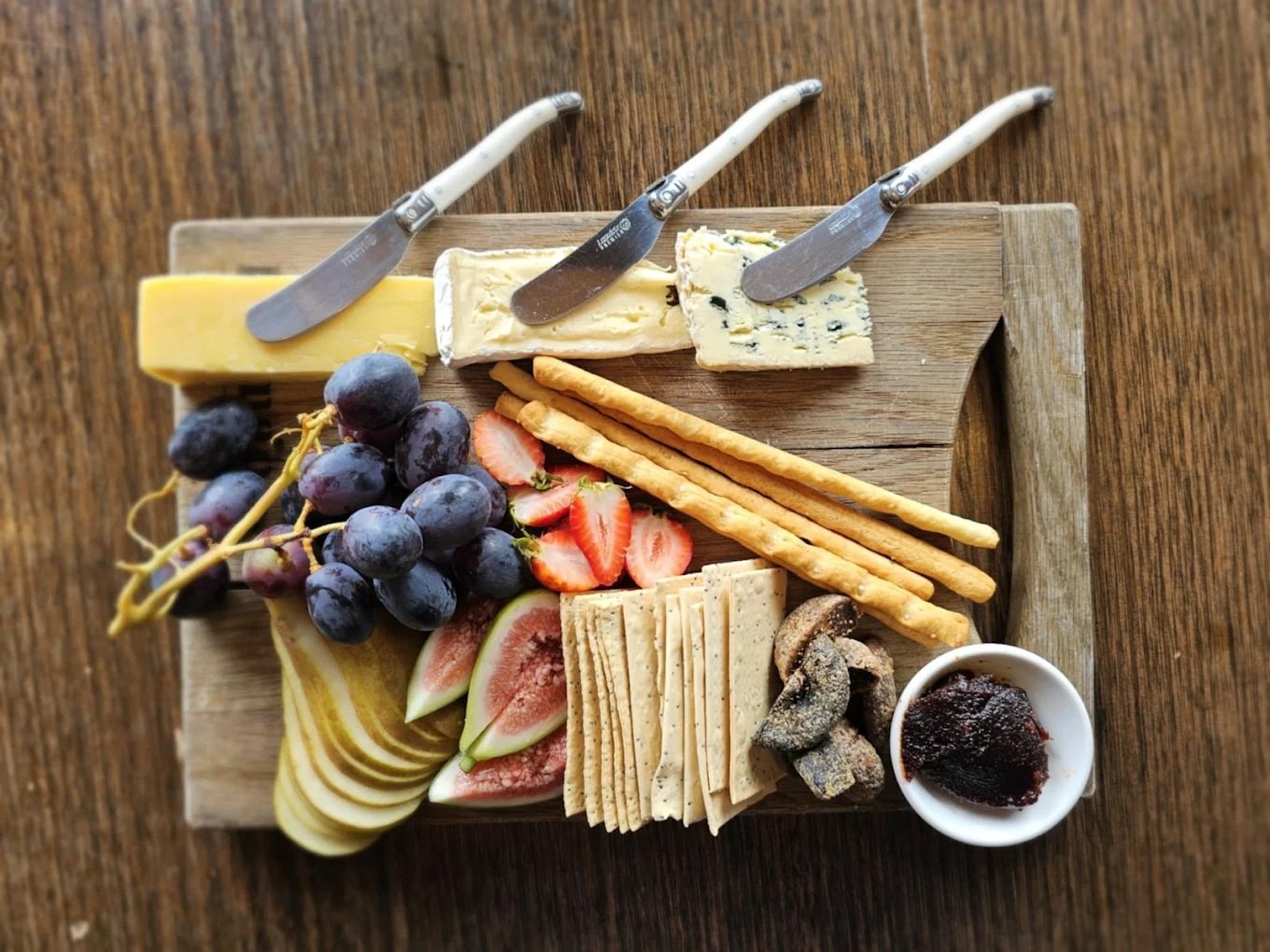 Cheese platter with grapes, strawberries, figs, sliced apples and crackers, served with a side of jam.