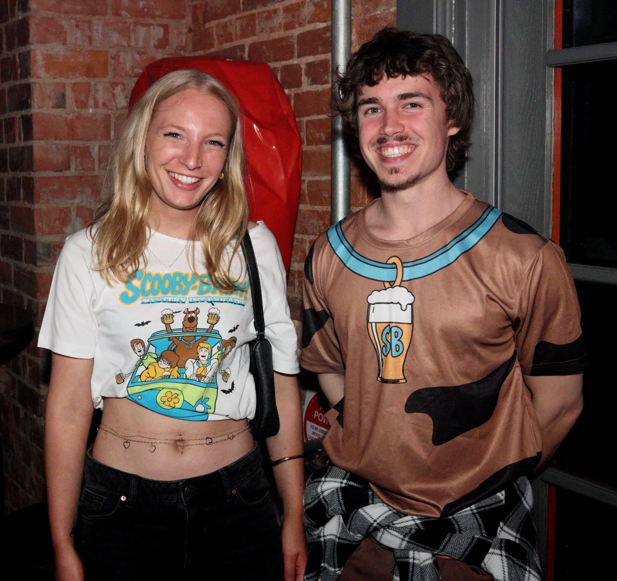 Young people dancing and socializing at a nightclub or party with colorful lights and laser effects, some wearing matching gray t-shirts with orange and green graphics.