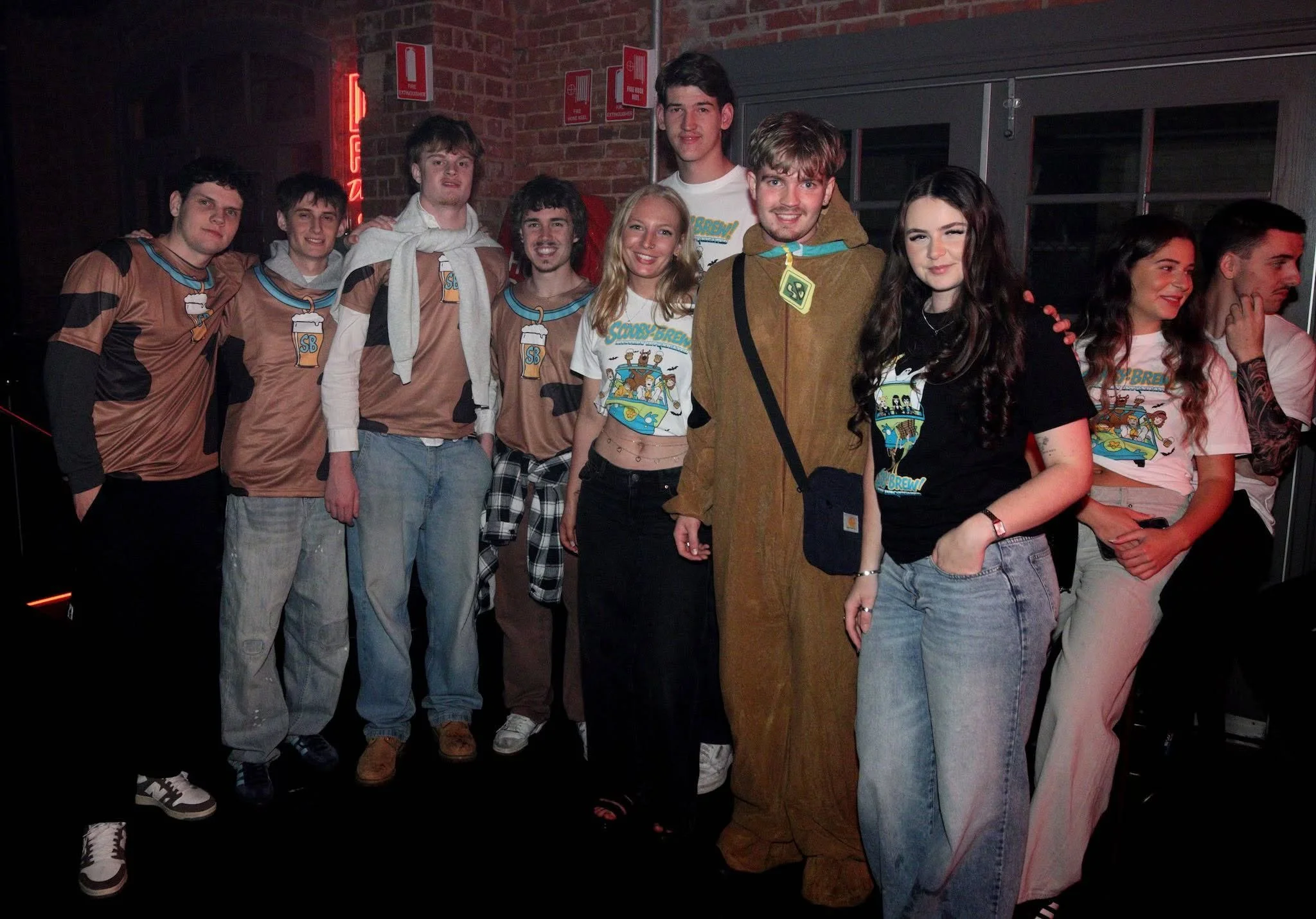 Group of people wearing colorful T-shirts in green, yellow, and white, posing together outside a building at night, some making gestures and smiling.