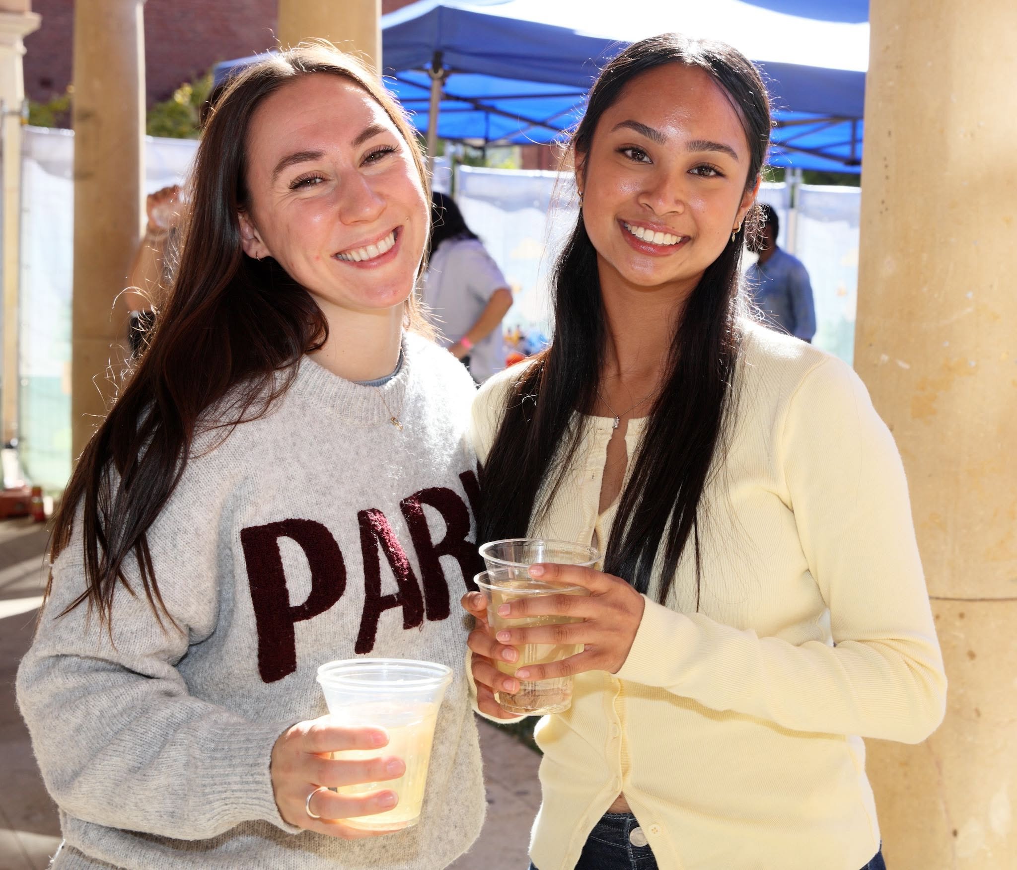 Group of young people in Hawaiian shirts and summer attire enjoying drinks at a sunny outdoor event with a booth selling beverages under a canopy.