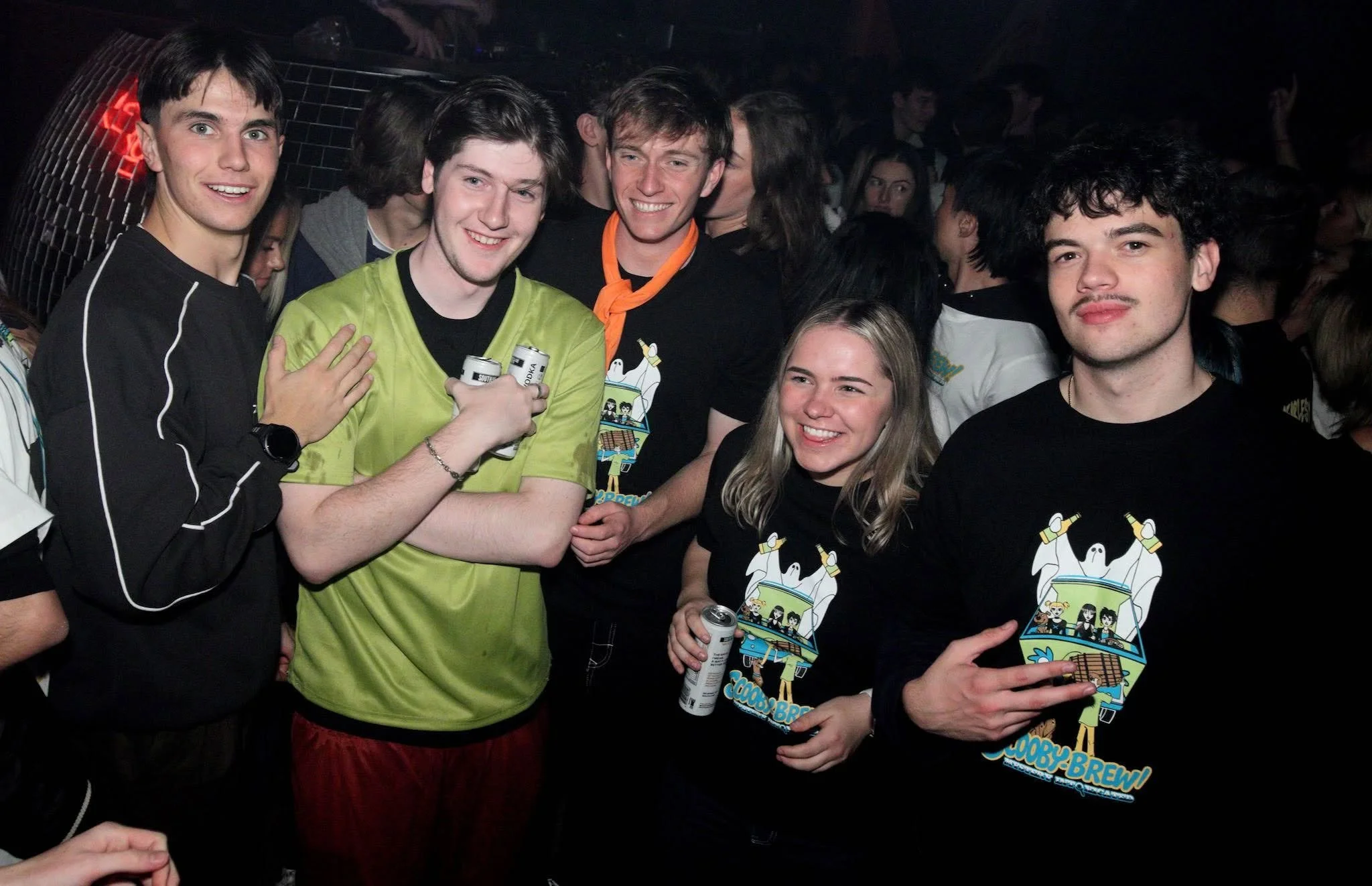 Four people wearing green Scrabble-themed T-shirts, spelling out 'AUES' across their shirts, at a social gathering, smiling and posing for the camera.