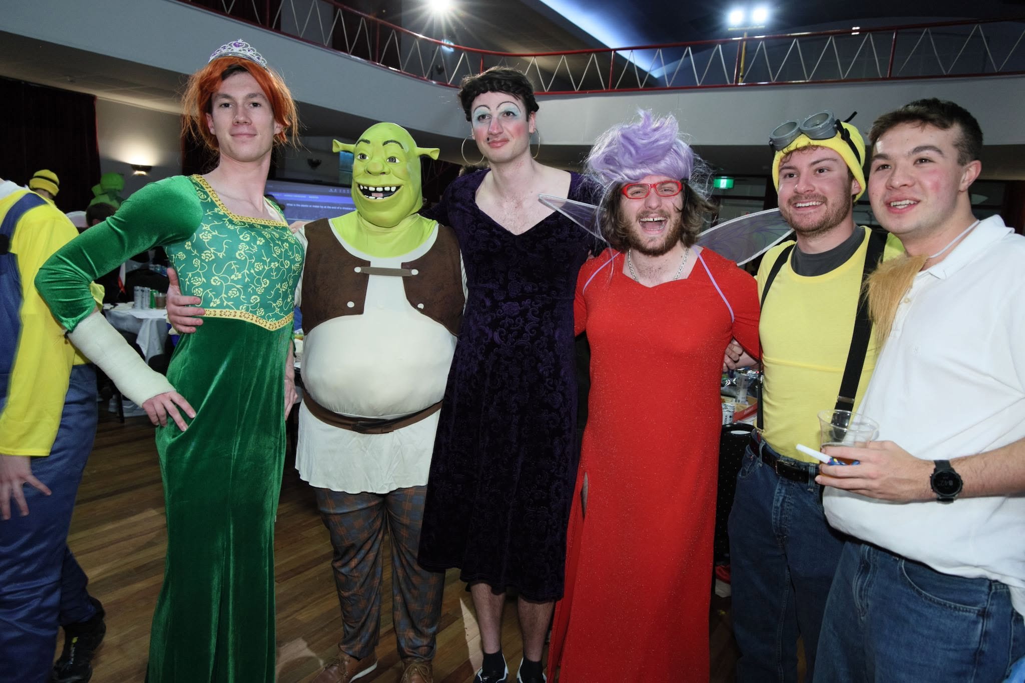 A large group of students at a decorated banquet hall celebrating, with many wearing yellow with 'Australia' on the back, some wearing masks, and in front, two students in red robes standing on tables with a broom and guitar, while audience members are clapping and smiling.