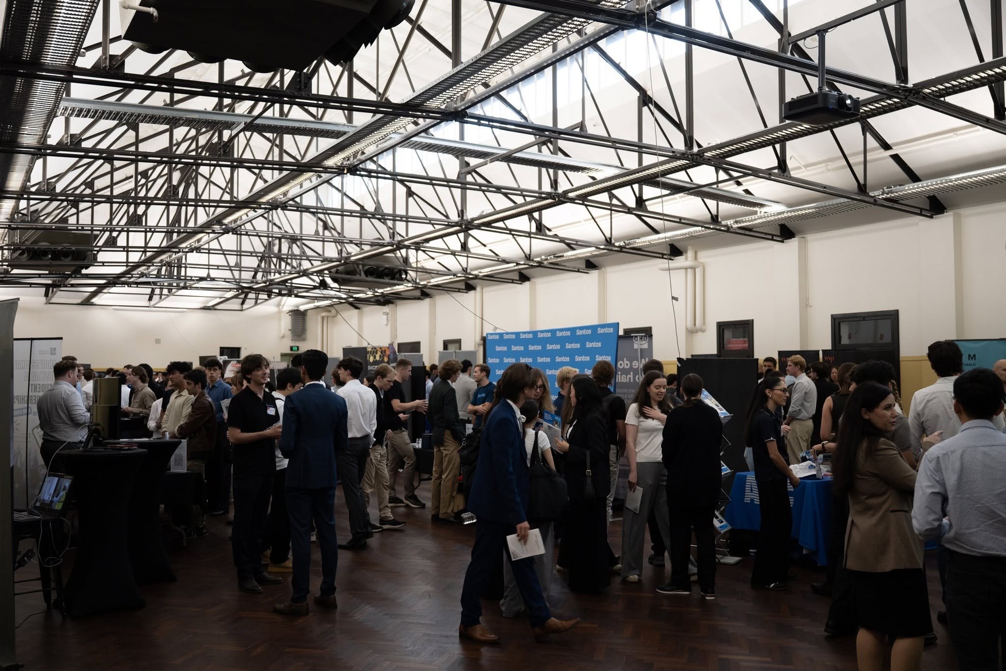 People attending a convention or expo, browsing and interacting at a long table with brochures and items, in a large indoor venue with displays and signs.