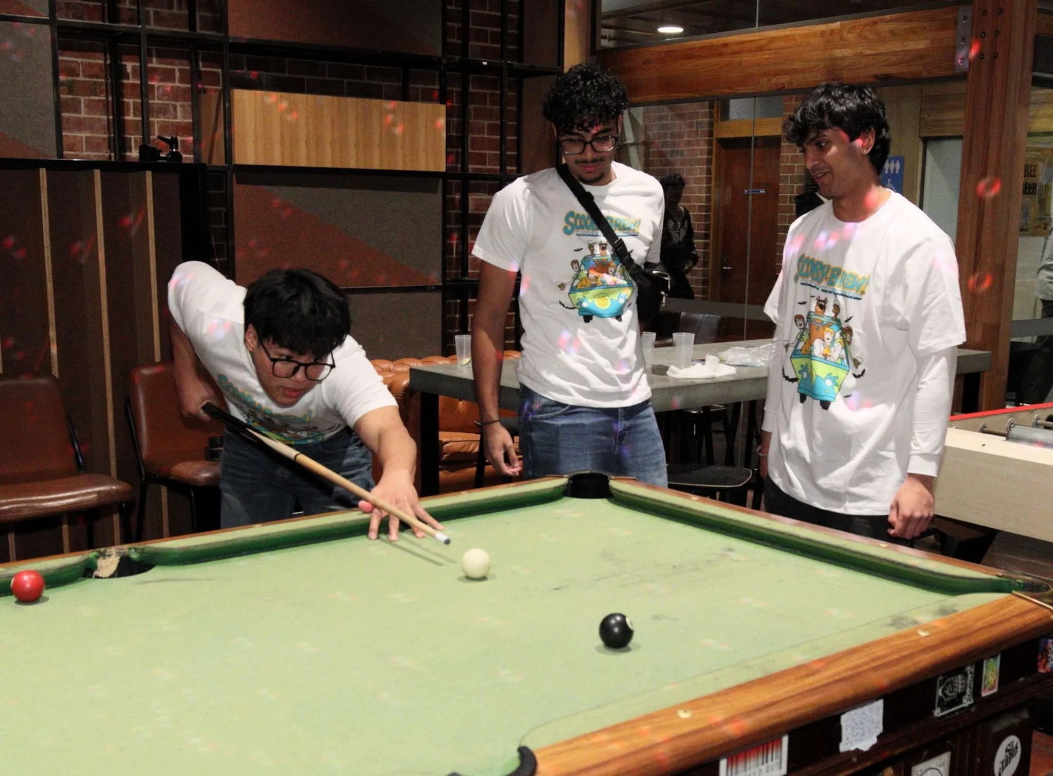 Group of young people posing and sitting in a lively indoor venue, some wearing matching event shirts, with colorful lighting and a bar in the background.