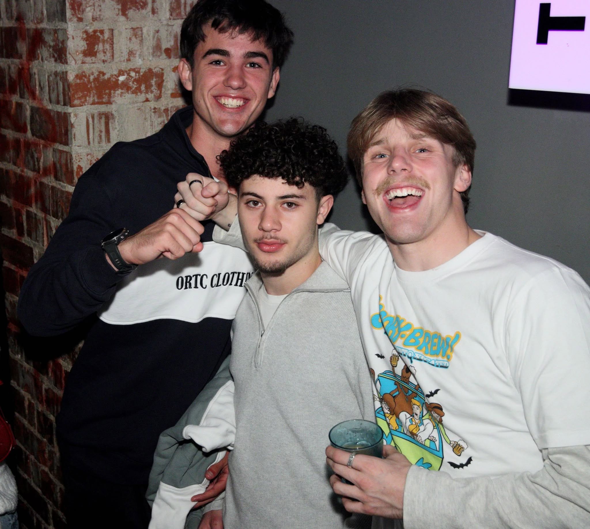 Group of five young adults smiling at a party or nightclub, wearing black team shirts with orange and red designs, with a lively crowd and DJ booth in the background.