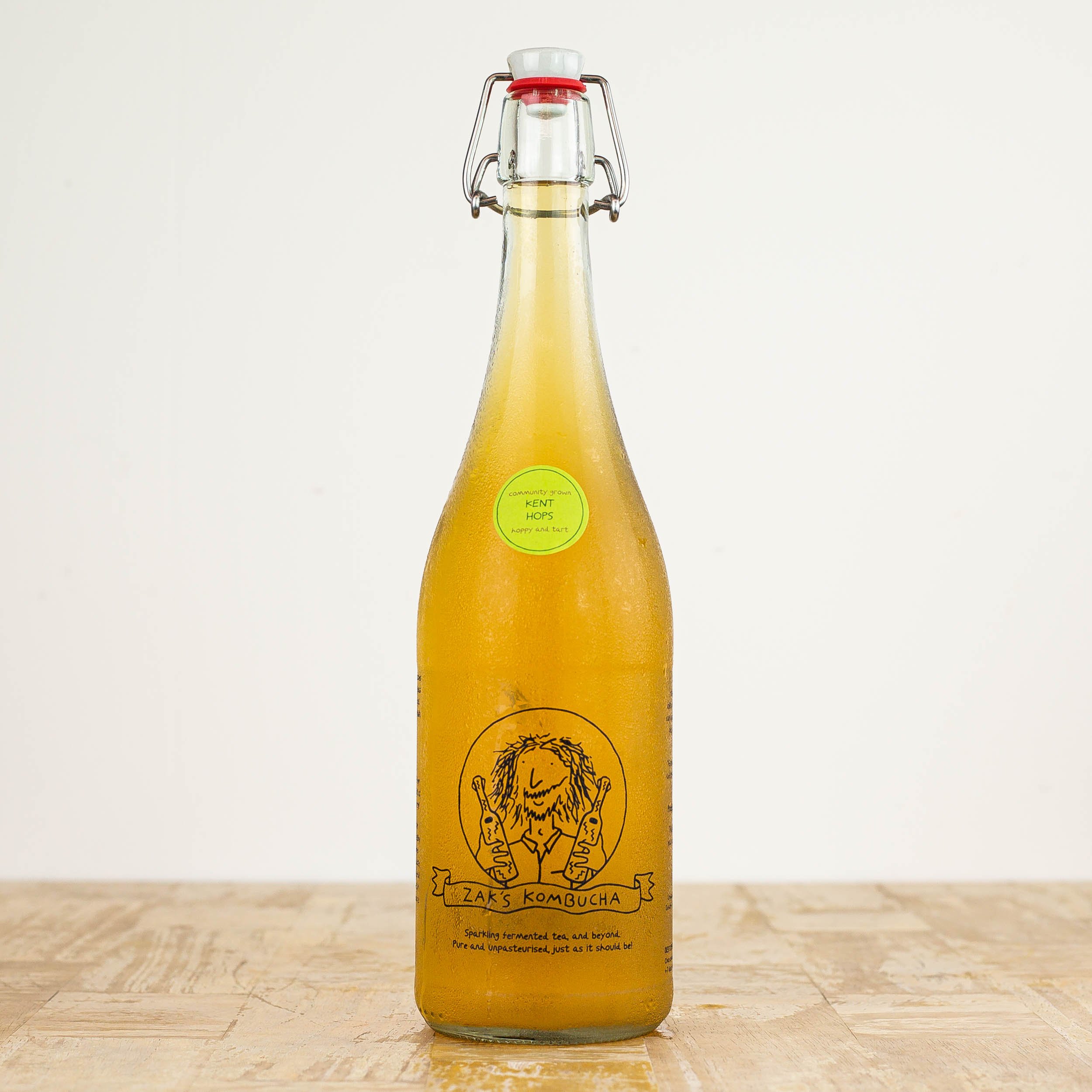 Single kent hops flavour zak's kombucha branded glass bottle, resting on a wooden table against a plain background.