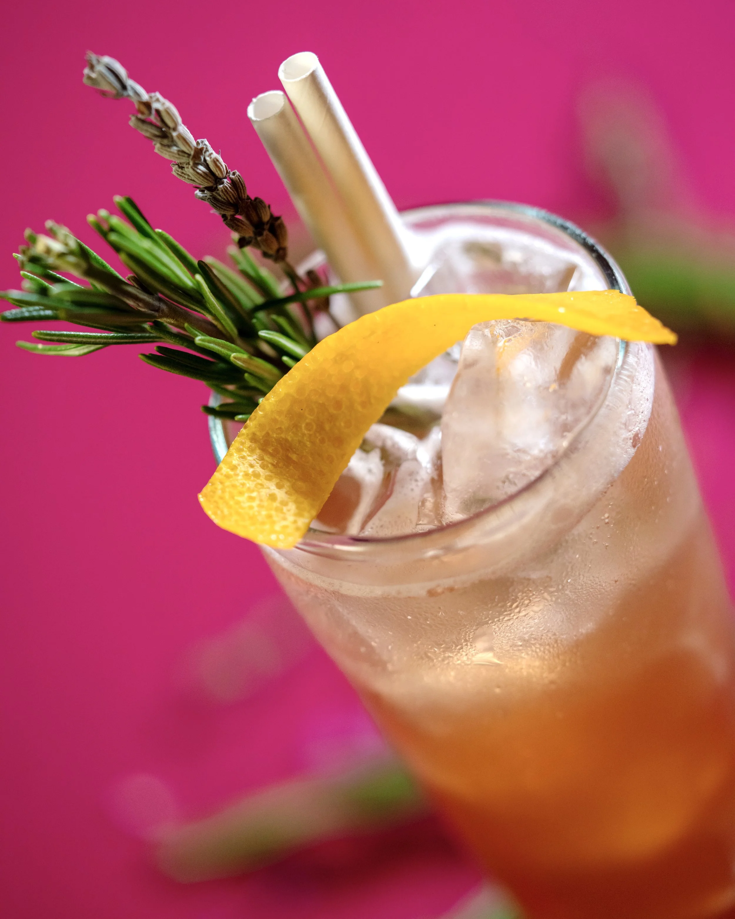 A close-up of an iced drink garnished with a lemon wedge, lavender sprig, and two white drinking straws against a pink background.