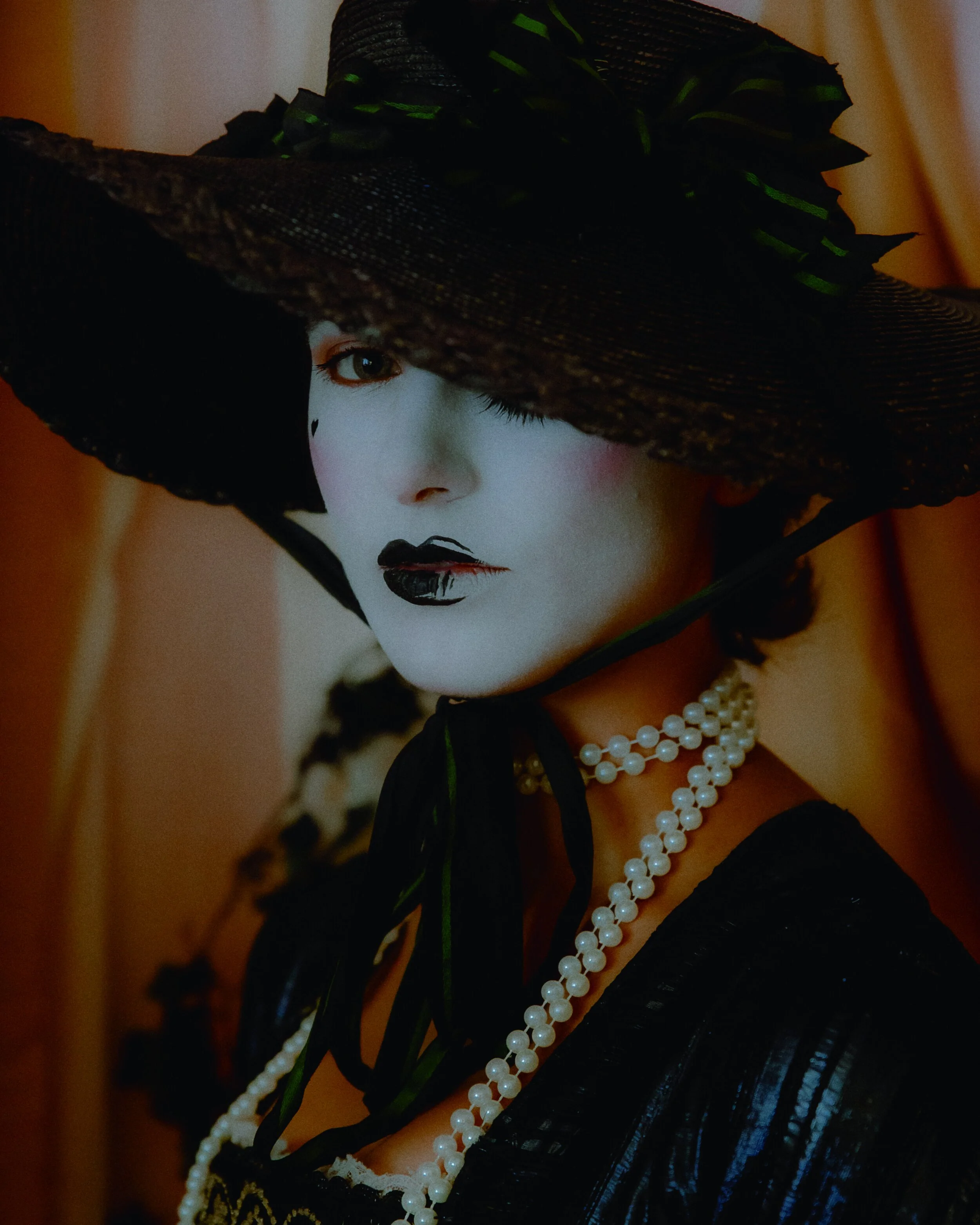 A woman with white face paint, black lipstick, and a large black hat with green accents. She is wearing layered pearl necklaces and a black dress, standing against a beige background.