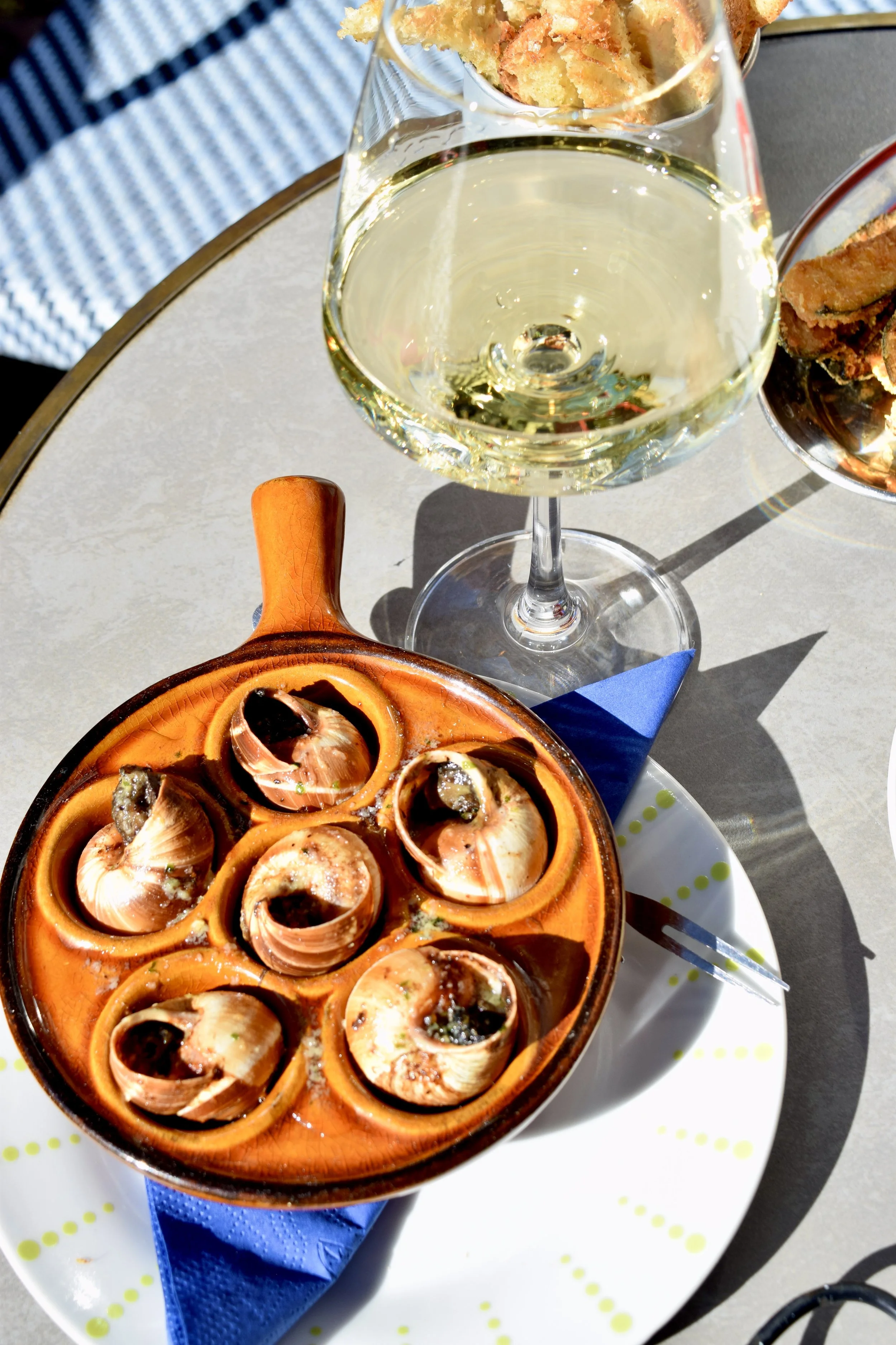A glass of white wine, a dish of baked escargots with garlic and herbs, and a bowl of crispy breadsticks on a patio table.