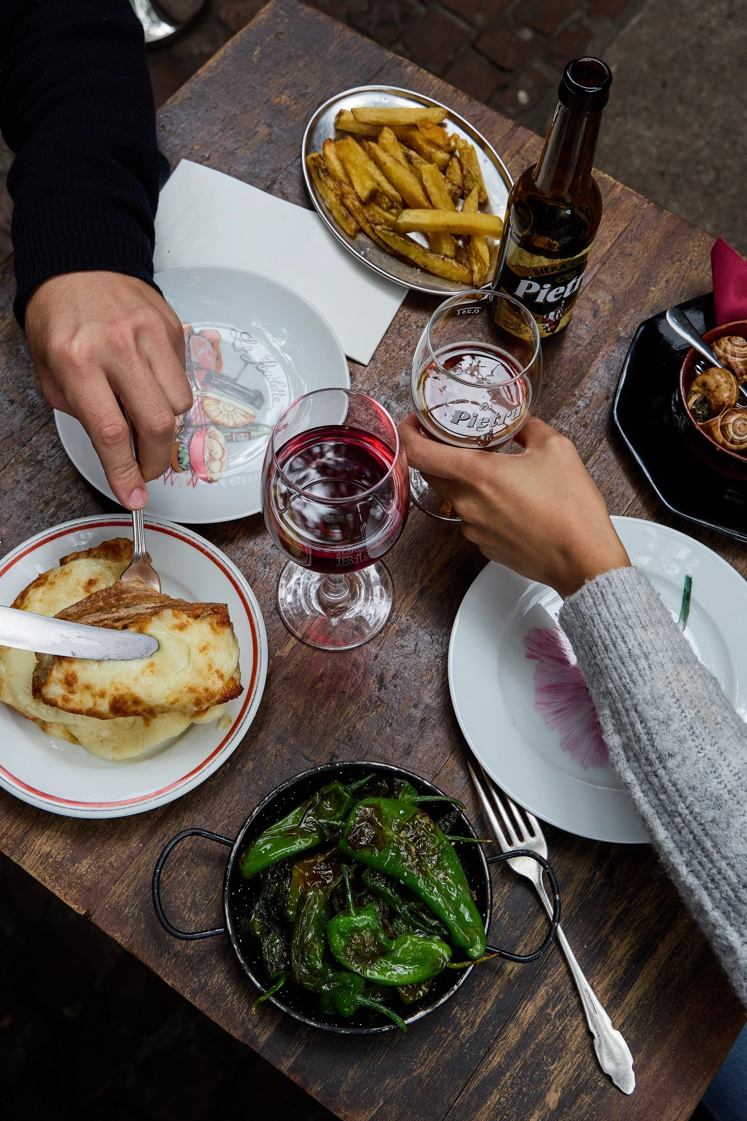 A table with two people celebrating with glasses of red and white wine, along with plates of food including baked dish with cheese, French fries, sautéed green peppers, and a bowl of snails, on a rustic wooden table.