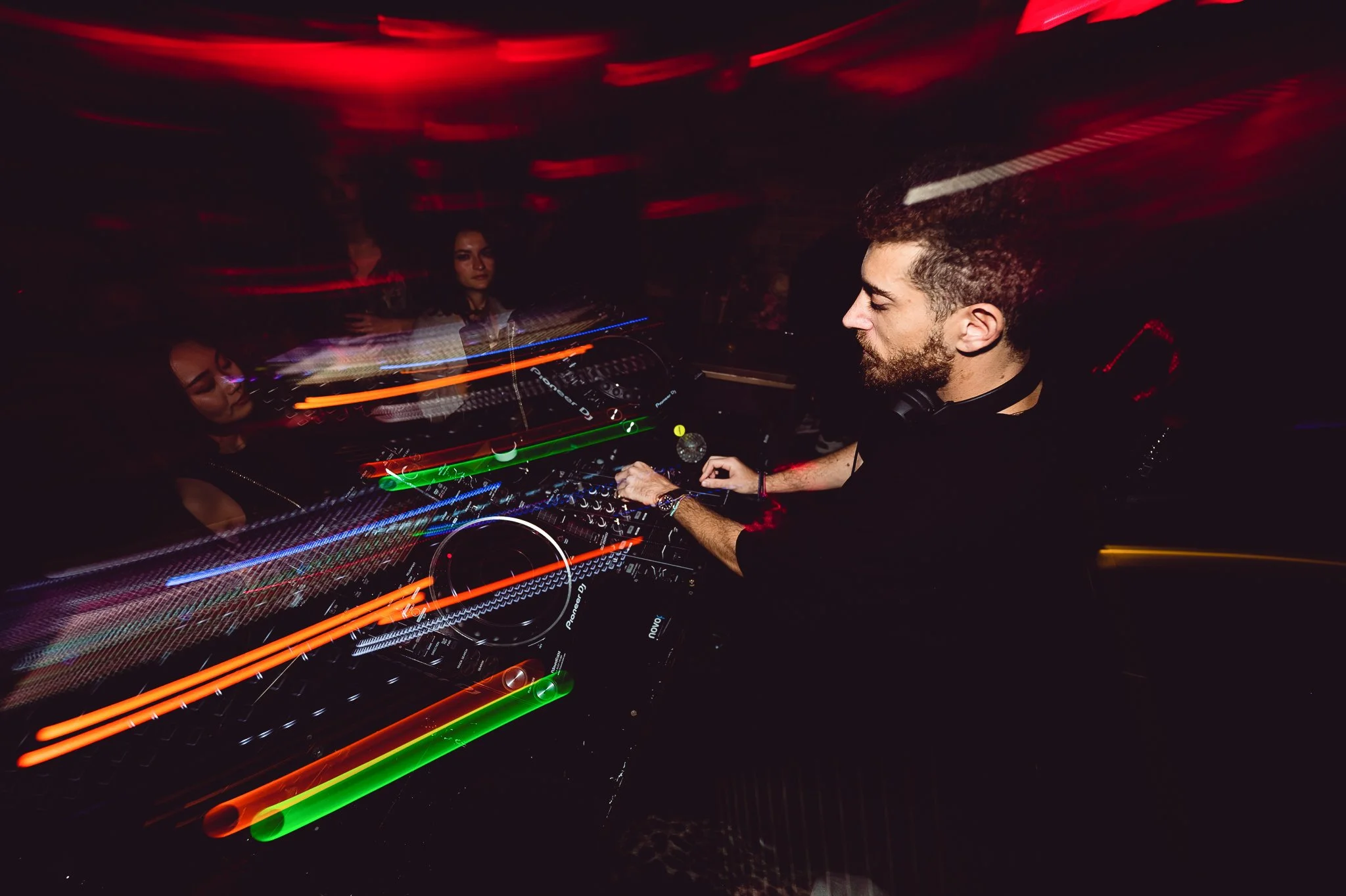 A DJ with a beard and headphones around his neck is performing at a nightclub, using a mixing console with colorful neon lights and motion blur effects in the background.