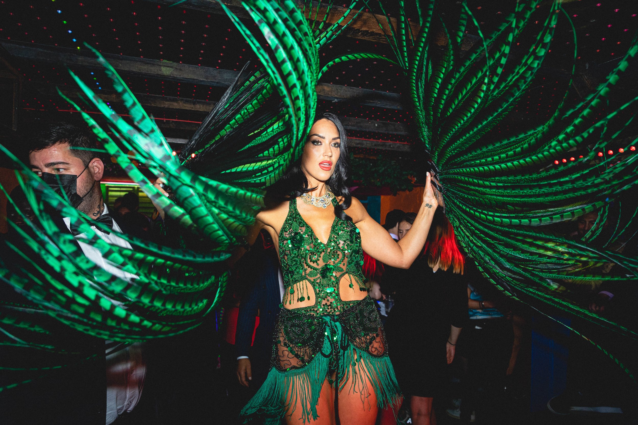 A woman in a green, ornate costume with large green wings at an indoor event or party. She has dark wavy hair and red lipstick. Other people are visible in the background, one man wearing a black mask and tuxedo.