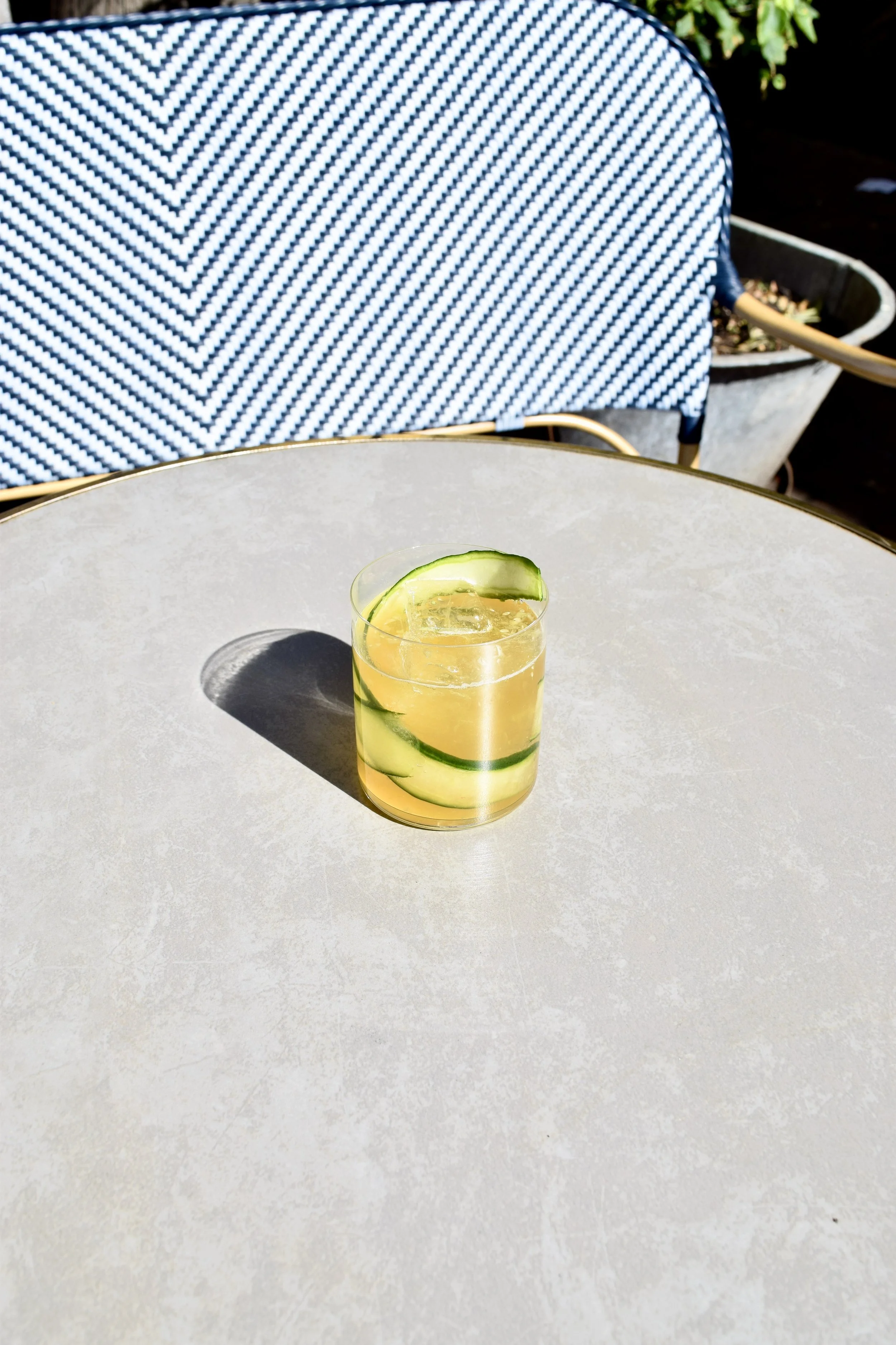 A glass with cucumber slices and a lemon wedge on a white outdoor table, casting a shadow in bright sunlight.