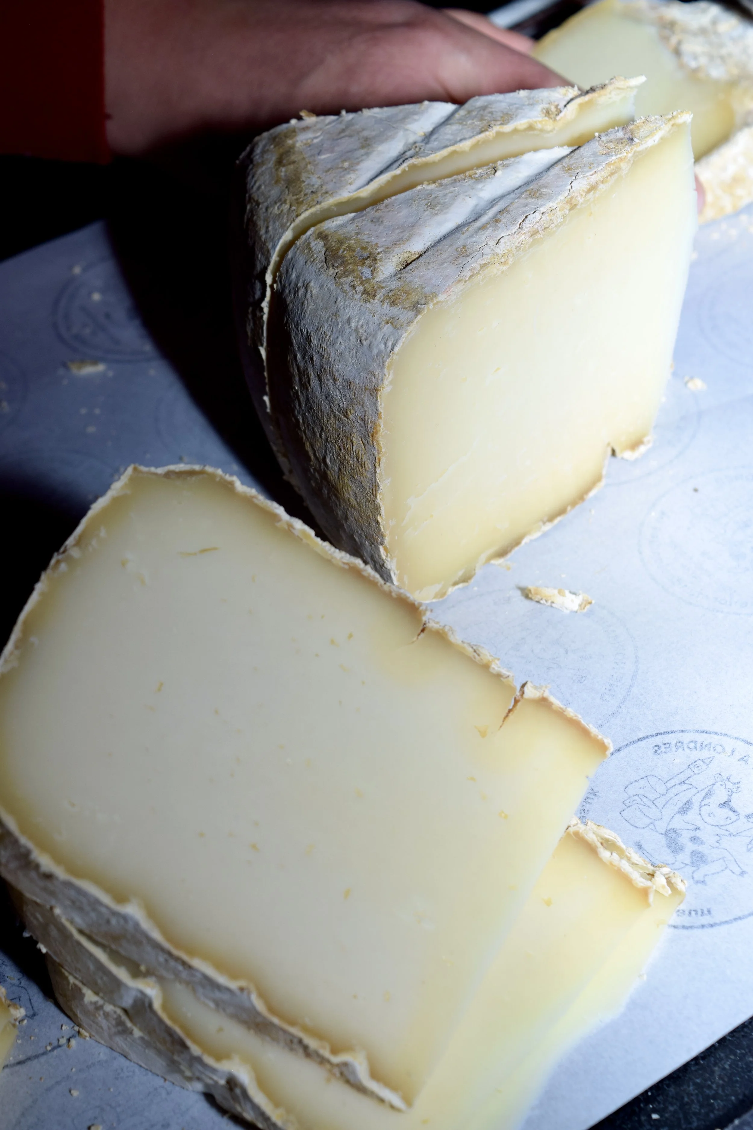 A block of aged cheese with a rind, partially sliced to reveal a firm, pale interior, resting on a piece of paper with a logo.