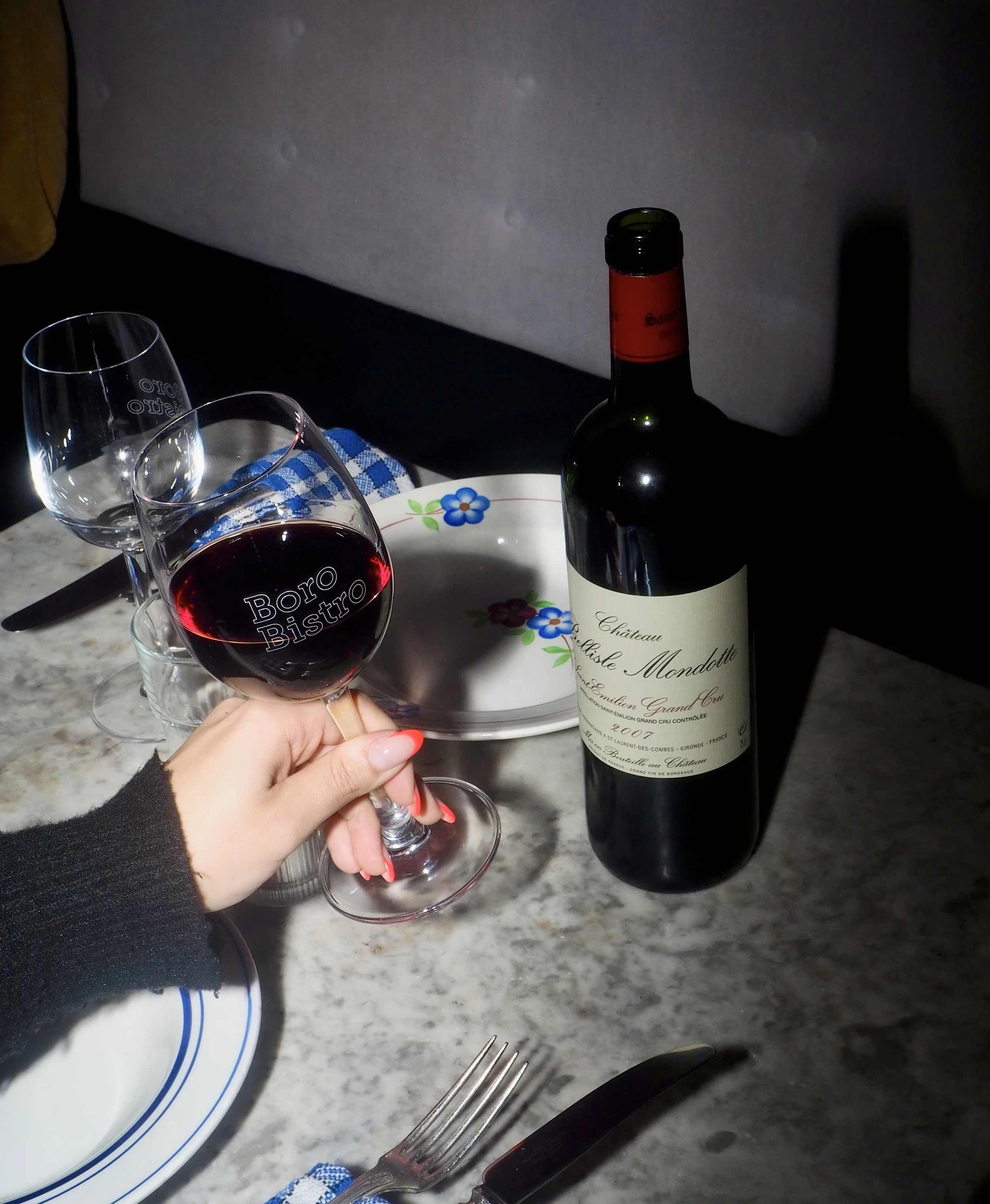 A person holding a glass of red wine at a dining table, with a bottle of red wine, a plate with floral design, two empty glasses, a fork, a knife, and a blue checkered napkin on a marble surface.