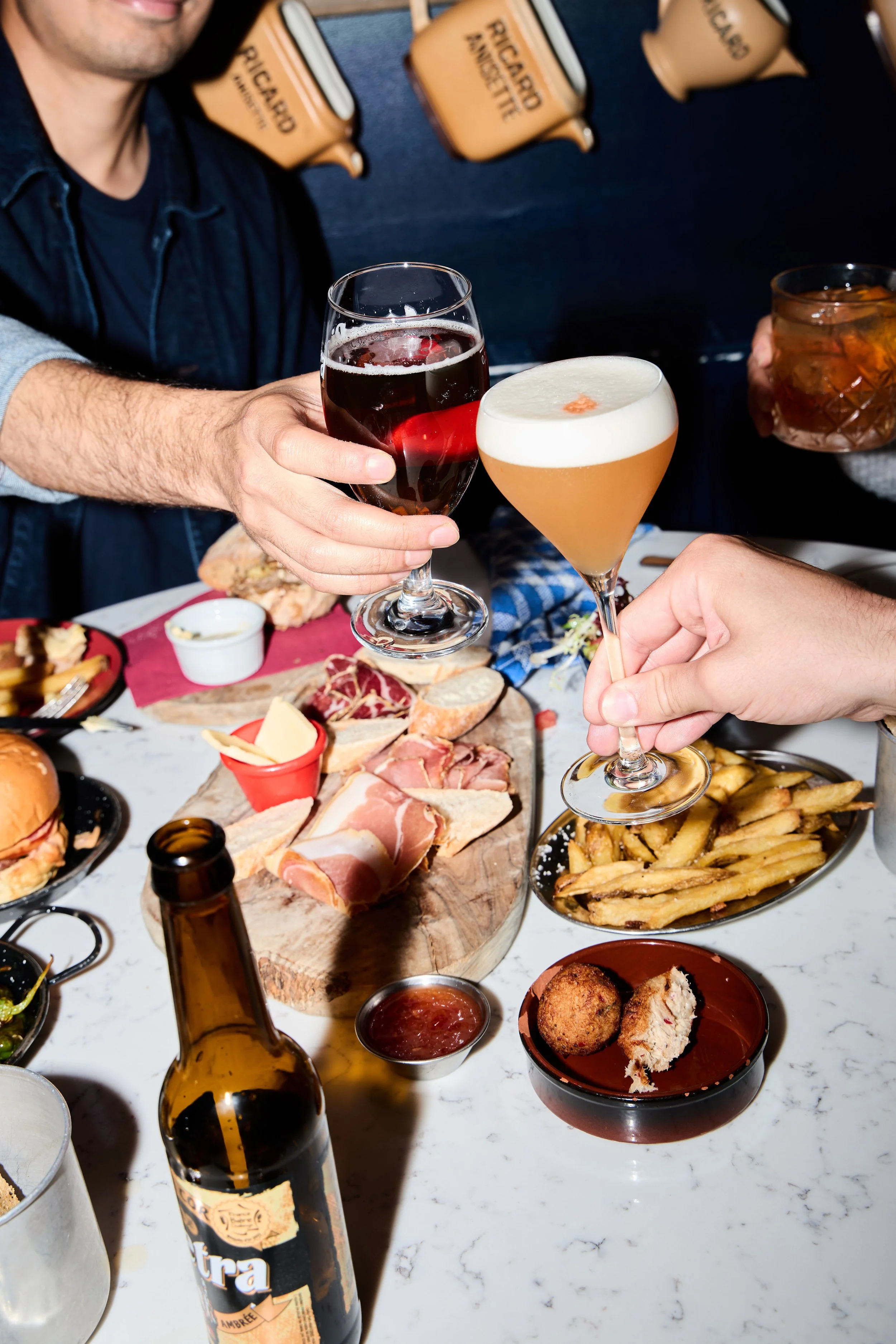 People raising glasses of red wine and cocktails in a toast over a table with food including French fries, charcuterie, bread, and fried appetizers.