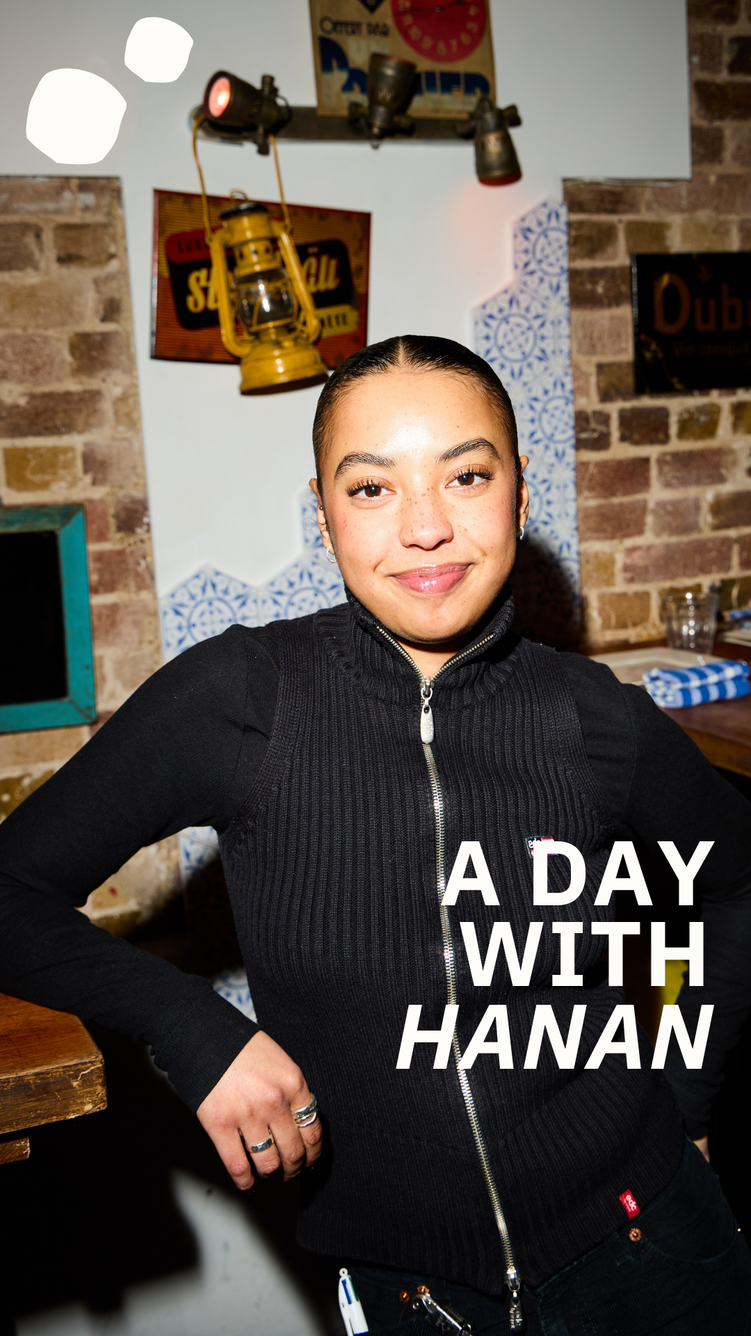 A woman with dark hair in a bun, smiling and standing against a rustic brick wall with decorative lamps and signs in the background, wearing a black zip-up sweater.