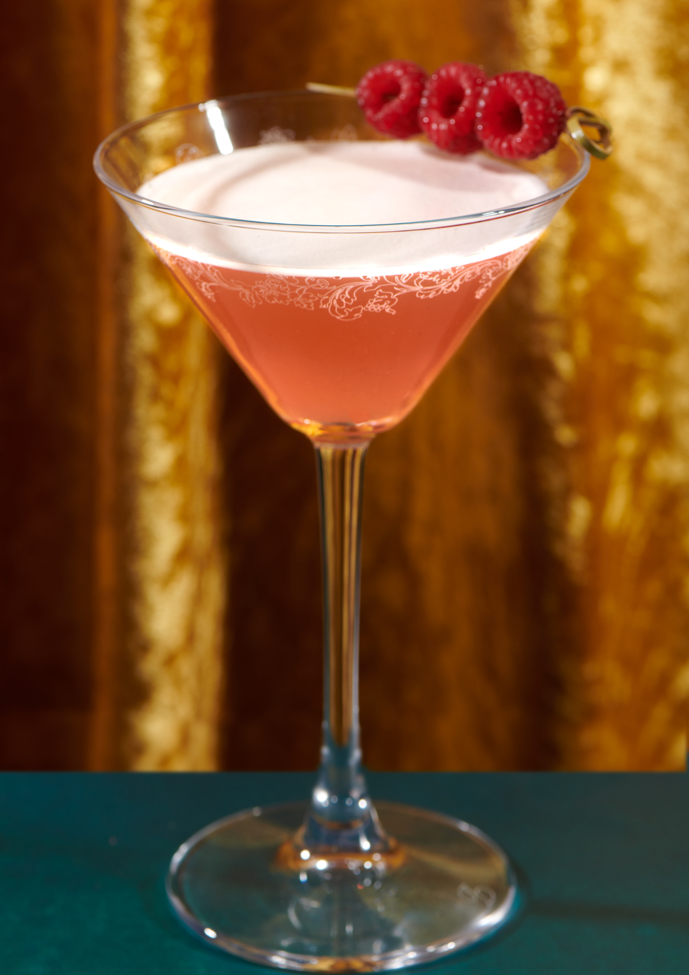 A pink cocktail in a martini glass garnished with three raspberries on a skewer, against a gold and brown background.
