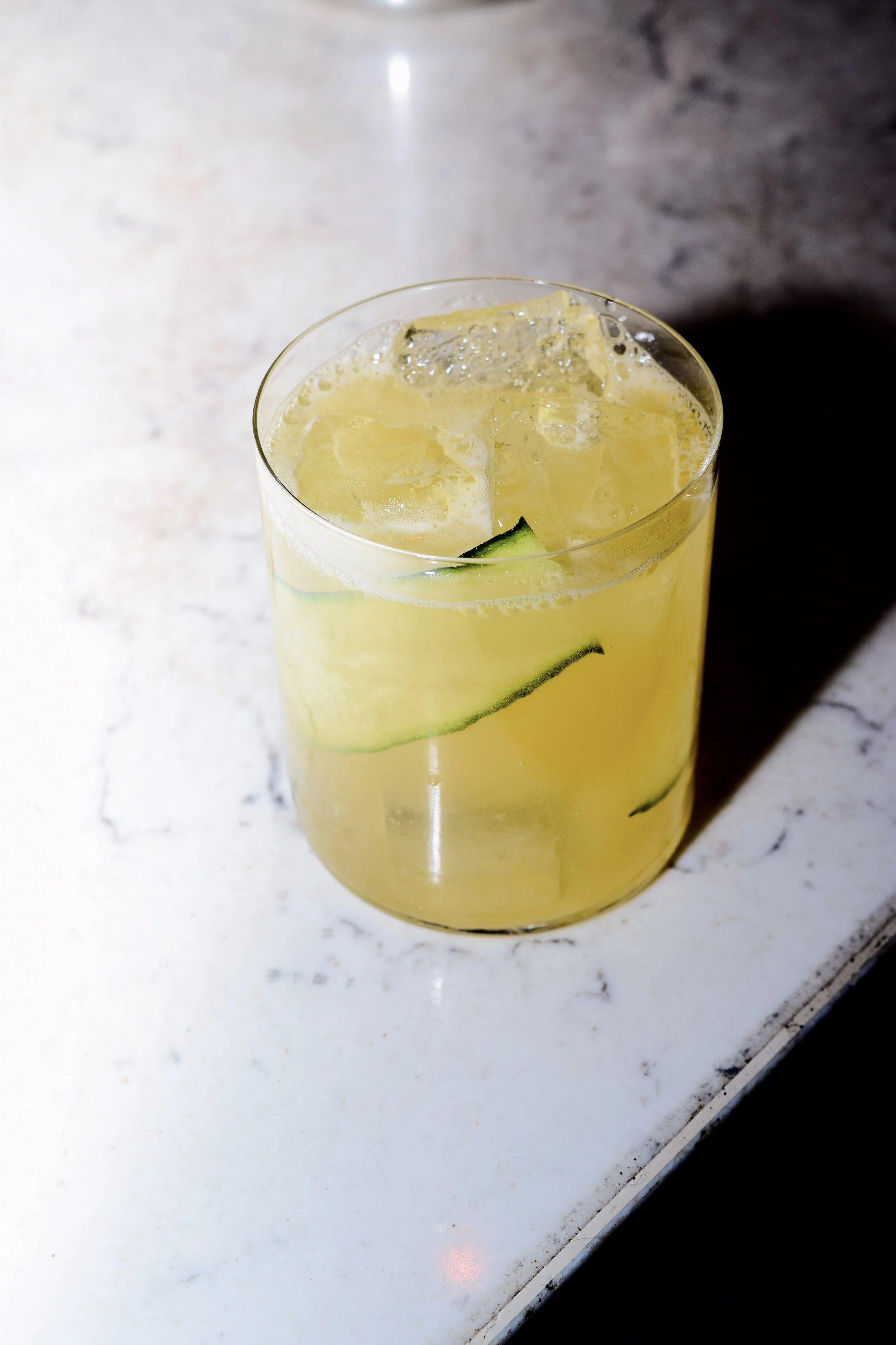 A transparent glass filled with a yellow drink, ice cubes, and cucumber slices on a light-colored marble surface.