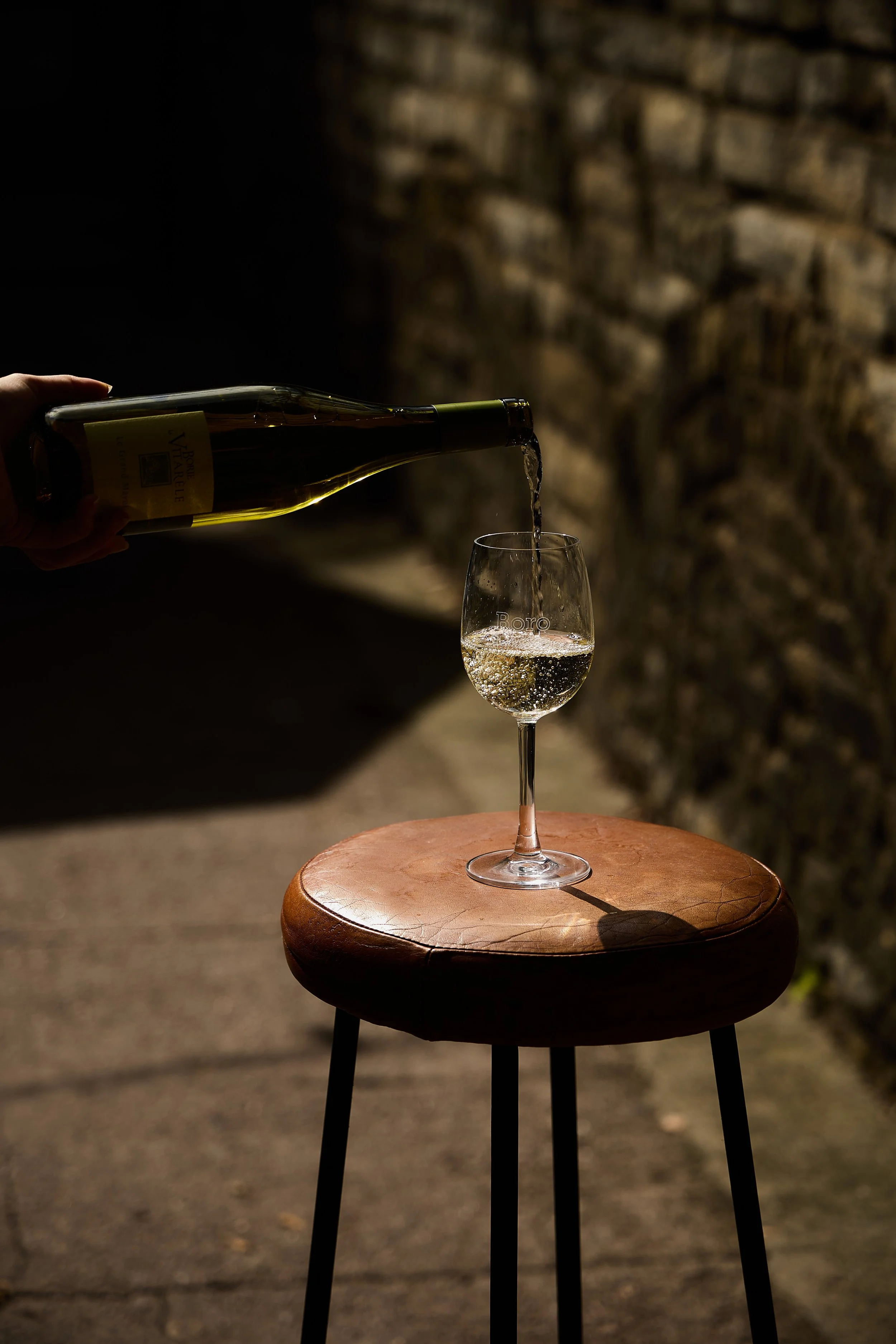 A hand pouring white wine into a glass on a wooden stool against a brick wall, with shadows and dim lighting.
