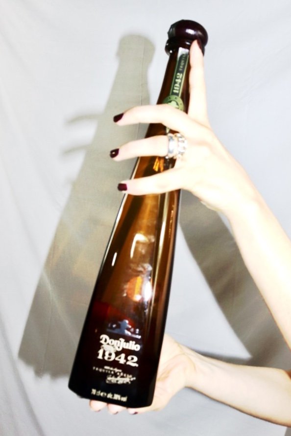 Person holding a brown glass bottle of Don Julio 1942 tequila, with fingers visible and long dark nail polish, in front of a plain background.