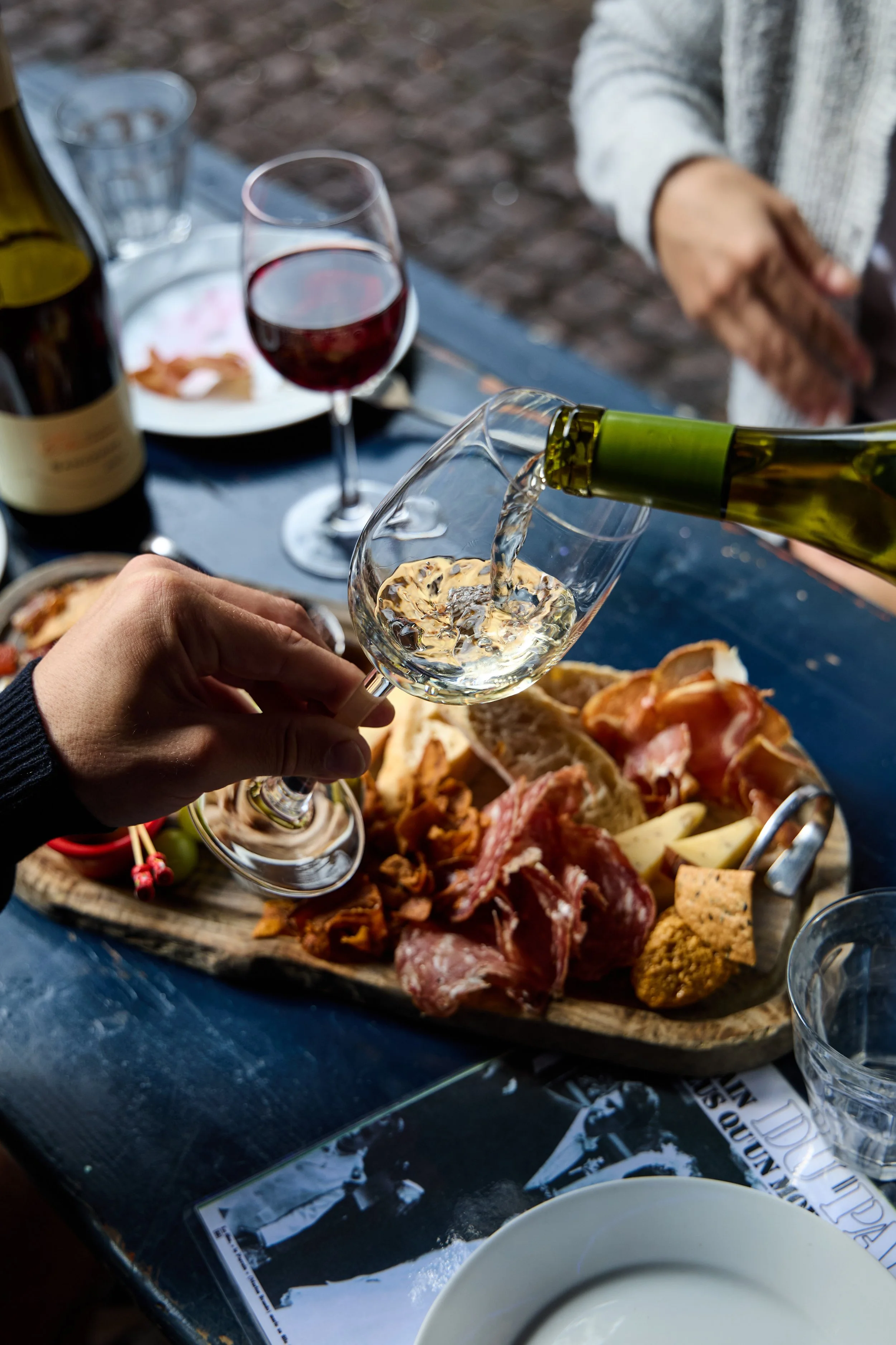 Person pouring white wine into a glass of red wine at a dining table with a charcuterie board of assorted meats and cheeses, and a glass of red wine in the background.
