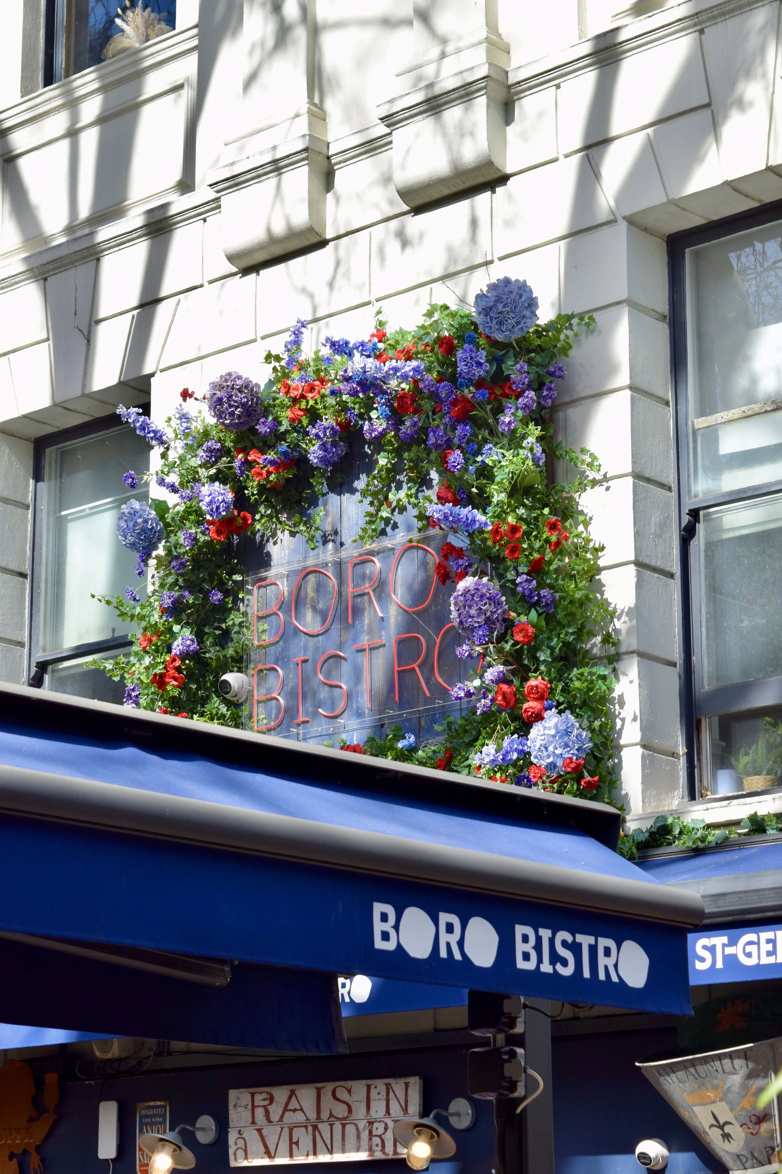 Building exterior with a neon sign reading 'BORO BISTRO' surrounded by colorful flowers, blue awning with 'BORO BISTRO' text, and a smaller sign showing 'RAISIN A VENDRE'.