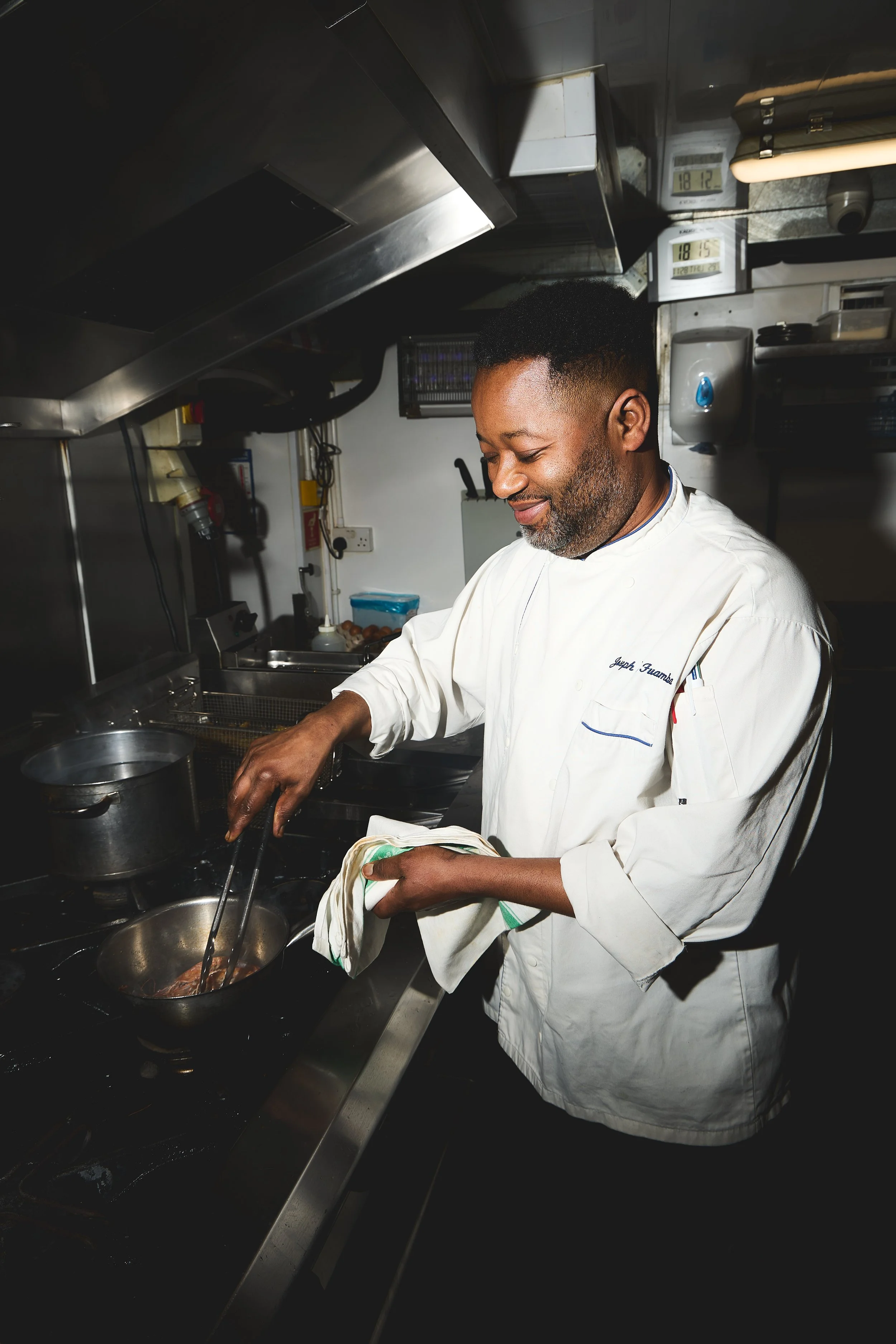 A chef in a white uniform smiles while cooking on a stove in a professional kitchen.