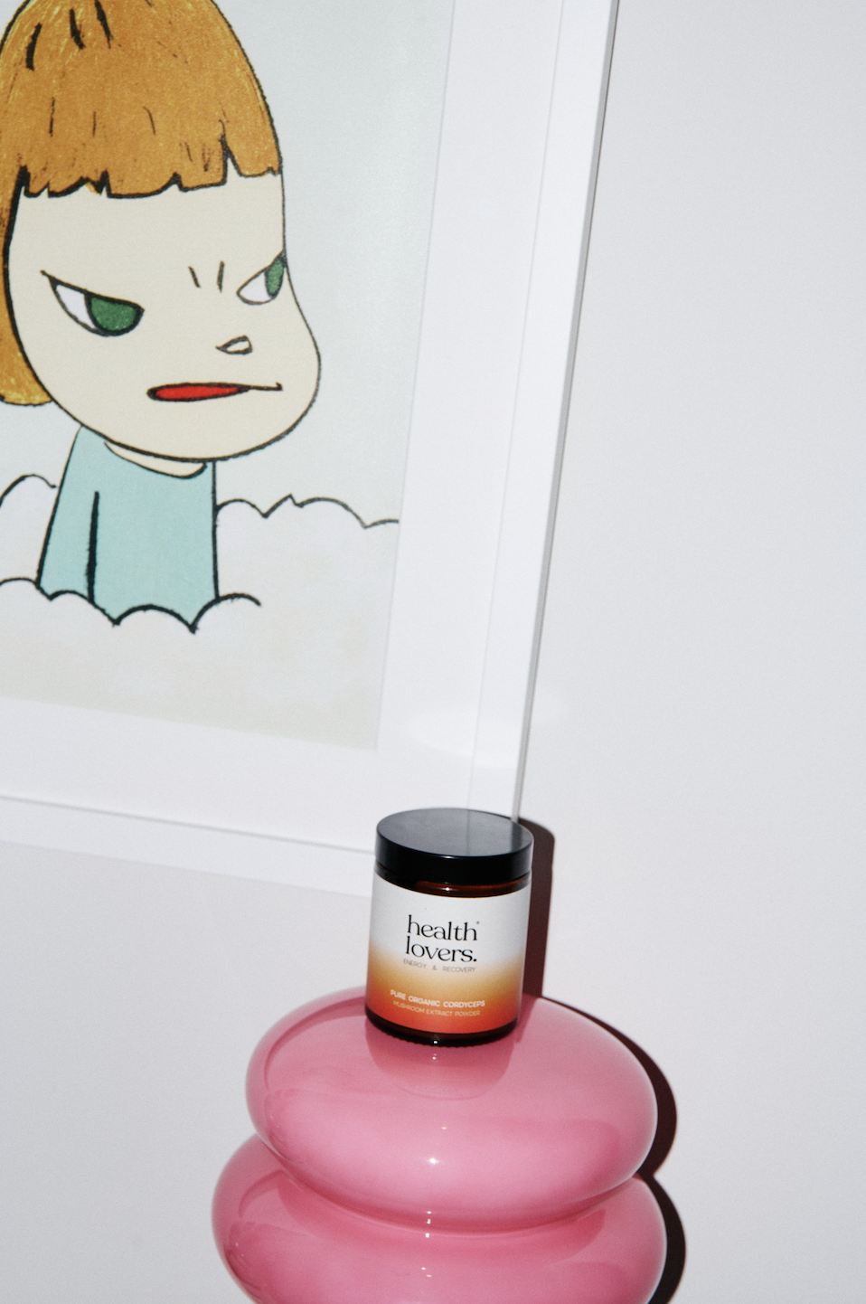 A cartoon character with a mischievous expression, brown hair, and a light blue shirt, depicted in a drawing or print on a wall. In front of it, there is a black container labeled 'health lovers' on a shiny pink, layered surface, with a thin rod extending from the container to the ceiling.