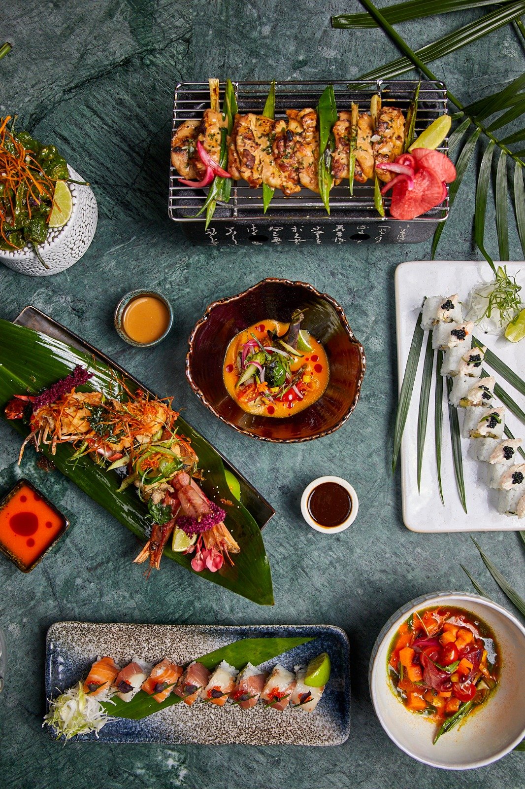 An assortment of Asian dishes laid out on a green surface, including grilled skewers, sushi rolls, a seafood platter, and various dipping sauces.