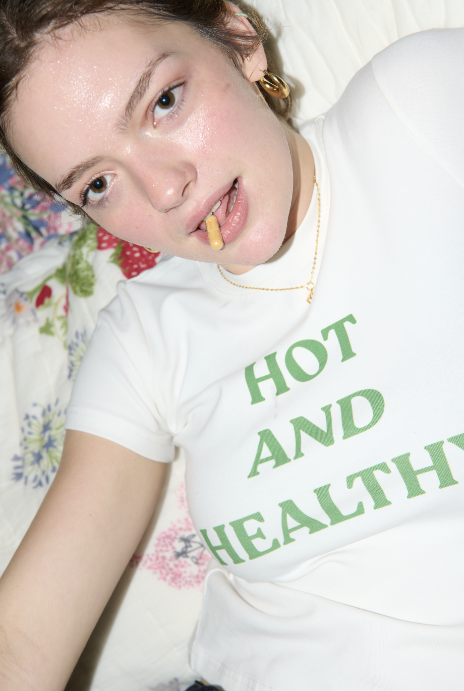 A young person with short brown hair, fair skin, and blue eyes lying on a bed with a floral quilt, wearing a white t-shirt with green text and a gold necklace, holding a pill between their teeth and an unlit cigarette in their mouth.