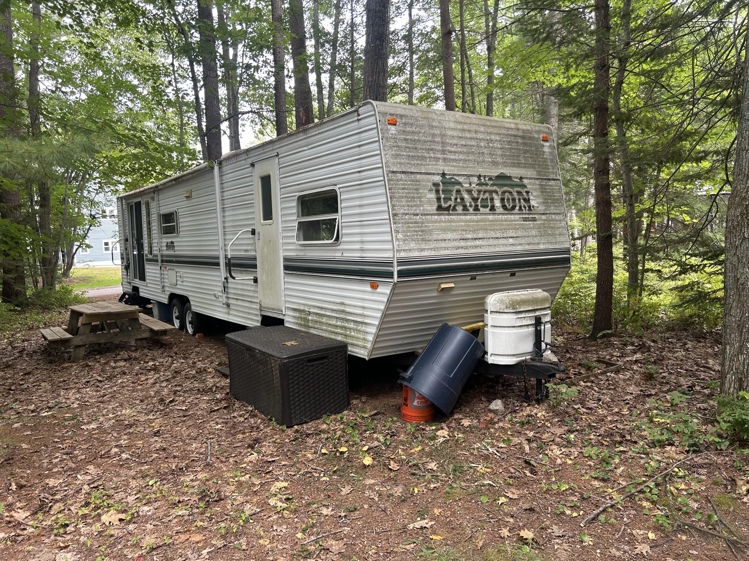 An old Layton camper in the woods with a wooden picnic table next to it