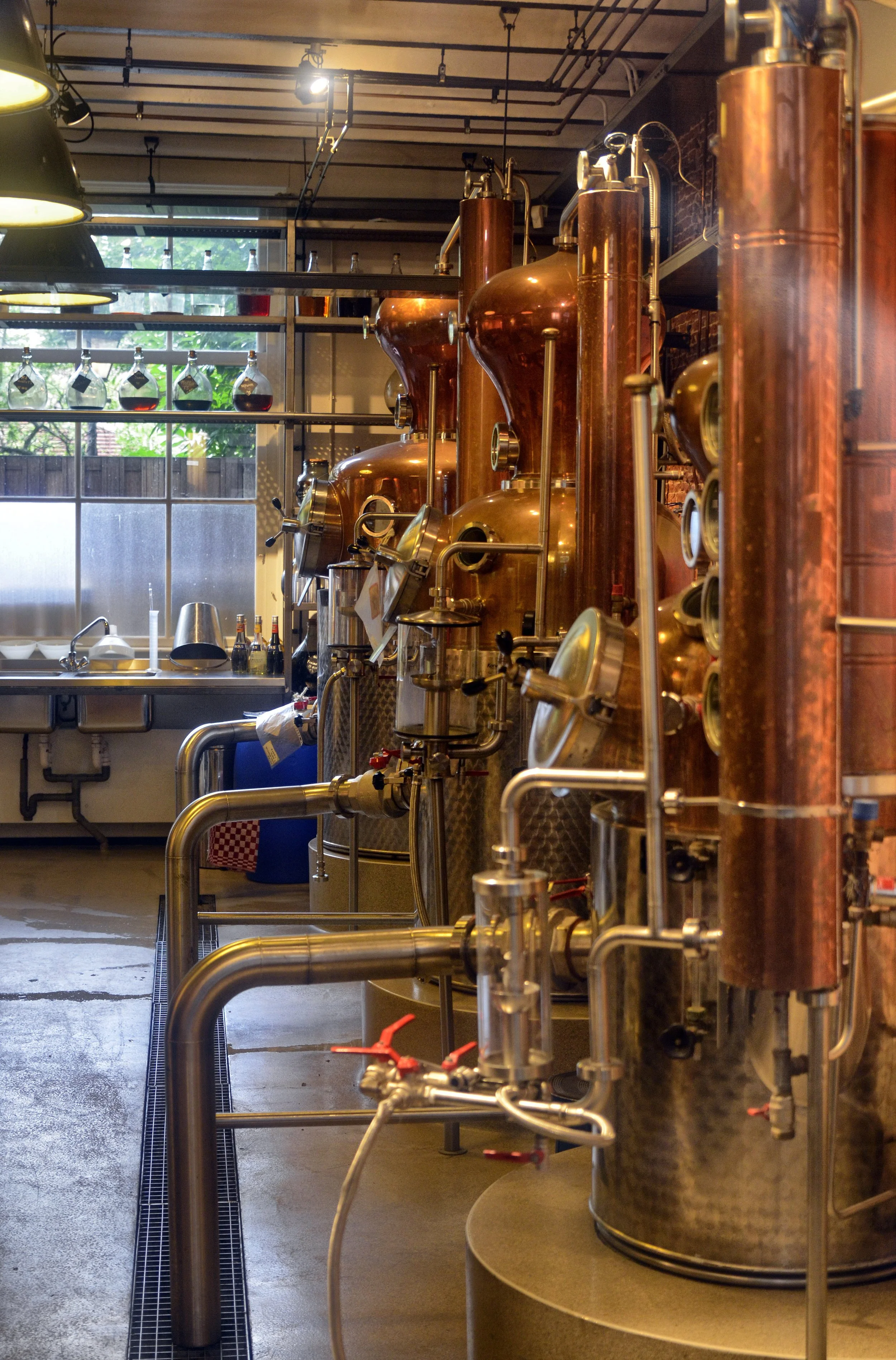 Copper distillation tanks in a distillery with a window and sink area in the background.
