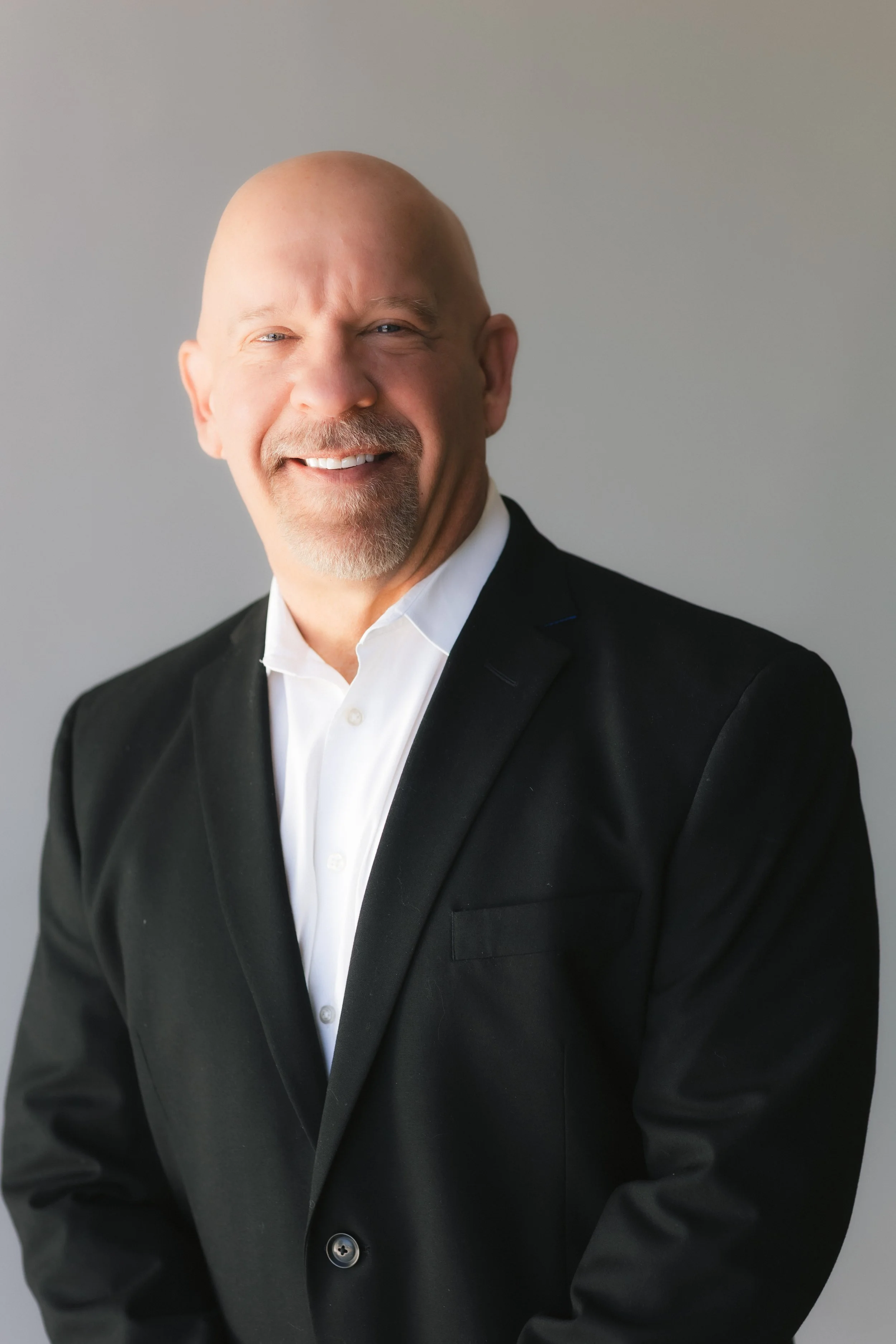 A bald man with a goatee smiling, wearing a black suit and white shirt, standing against a plain background.