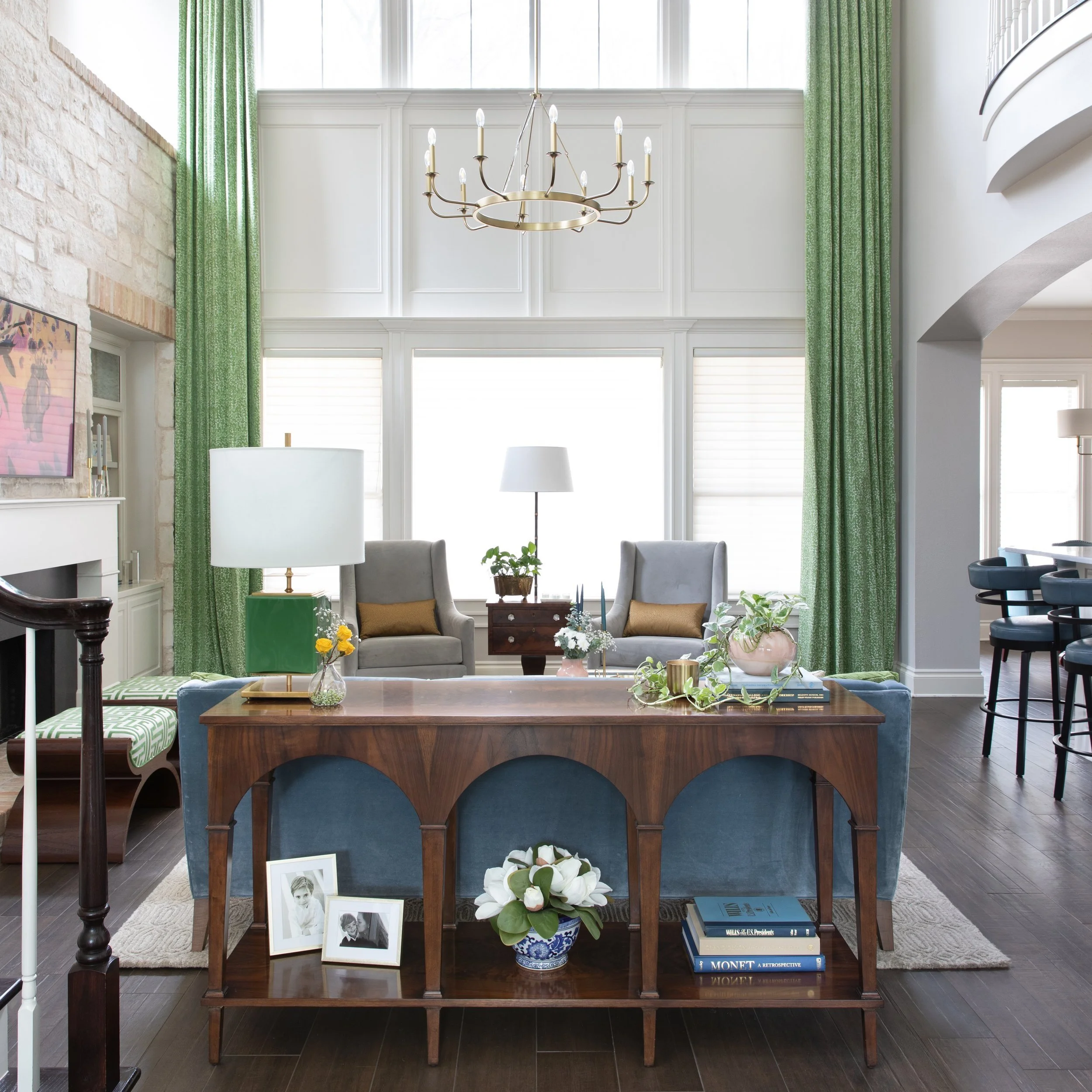 Living room seating area with stone accent wall, console table behind the couch, and a chandelier with tall green curtains open to reveal clerestory windows
