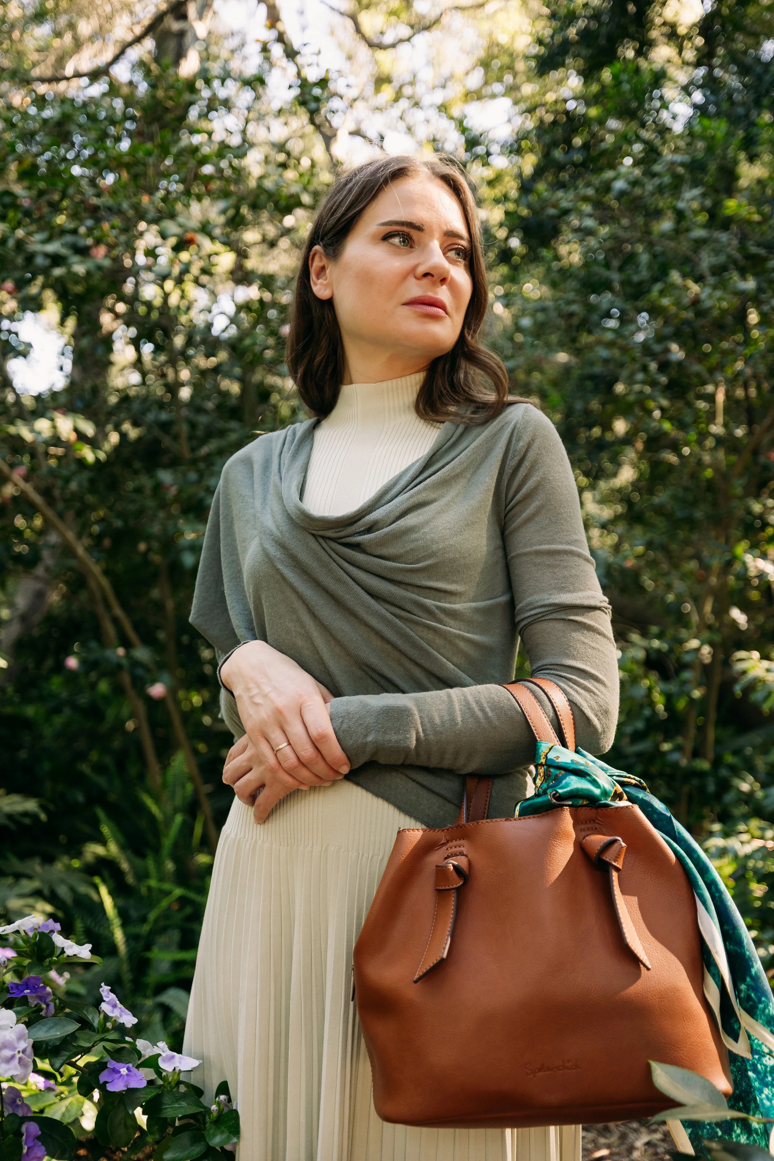 A woman with brown hair stands outdoors among greenery and flowers, wearing a cream turtleneck and an olive green cardigan, holding a large brown handbag with a colorful scarf tied to it.