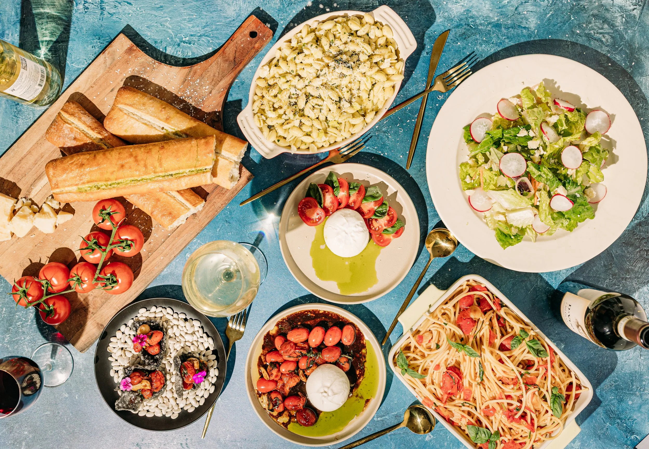 A table filled with various Italian dishes including a salad, pasta, bread, cherry tomatoes, and wine.
