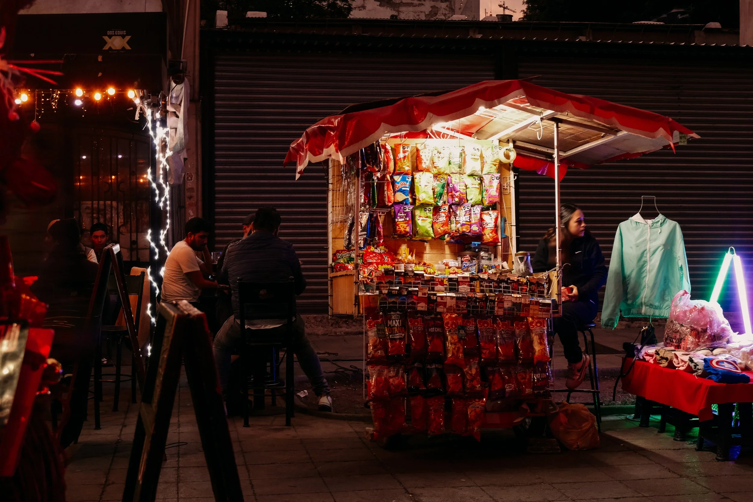 Nighttime street scene with a small snack stand lit up, showcasing bags of chips and candies, with people sitting nearby and closed shop shutters in the background.