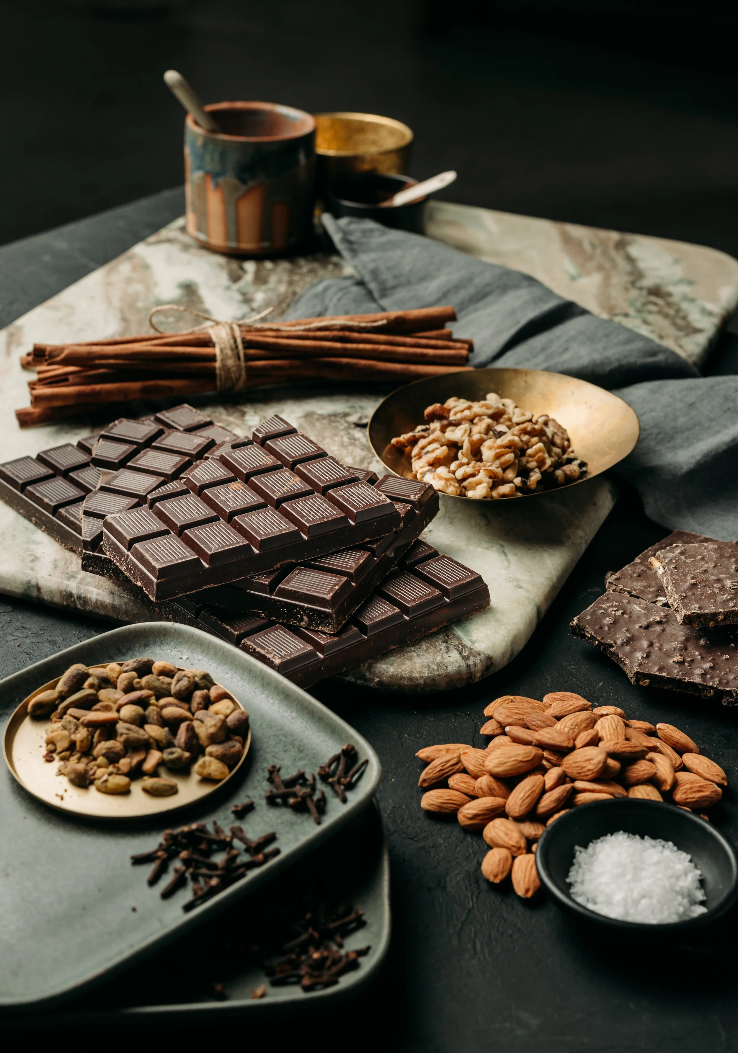 Assorted chocolate bars, almonds, sea salt, and baking ingredients on a slab and bowls, with a rustic backdrop.