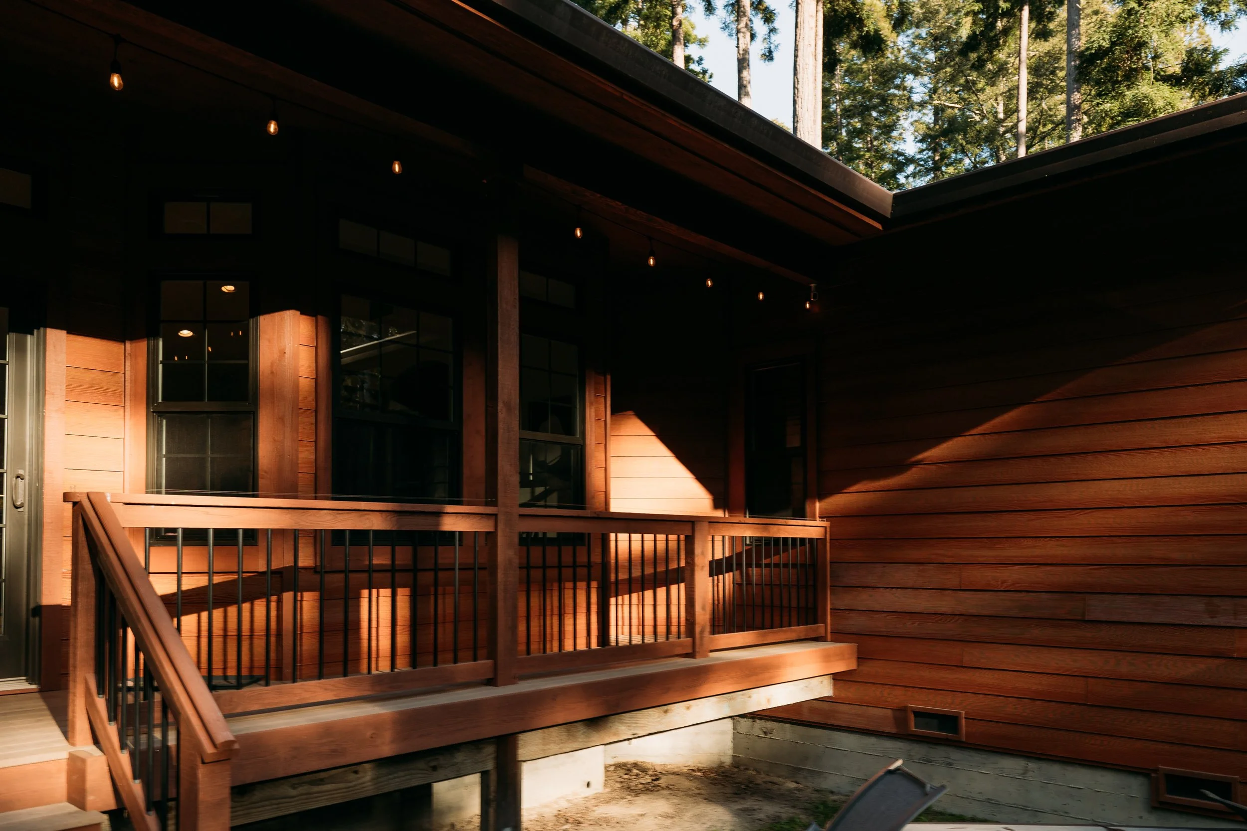Exterior of a wooden house with a small balcony, sunlight casting shadows on the wooden siding, with trees visible in the background.