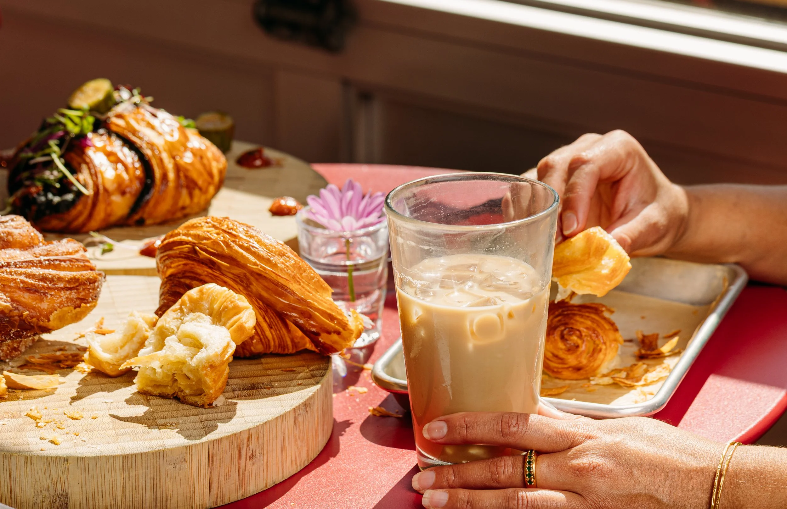 A breakfast with pastries on a wooden cutting board and a glass of iced coffee held by a person wearing rings, set on a red table with a pink flower in a small glass vase.