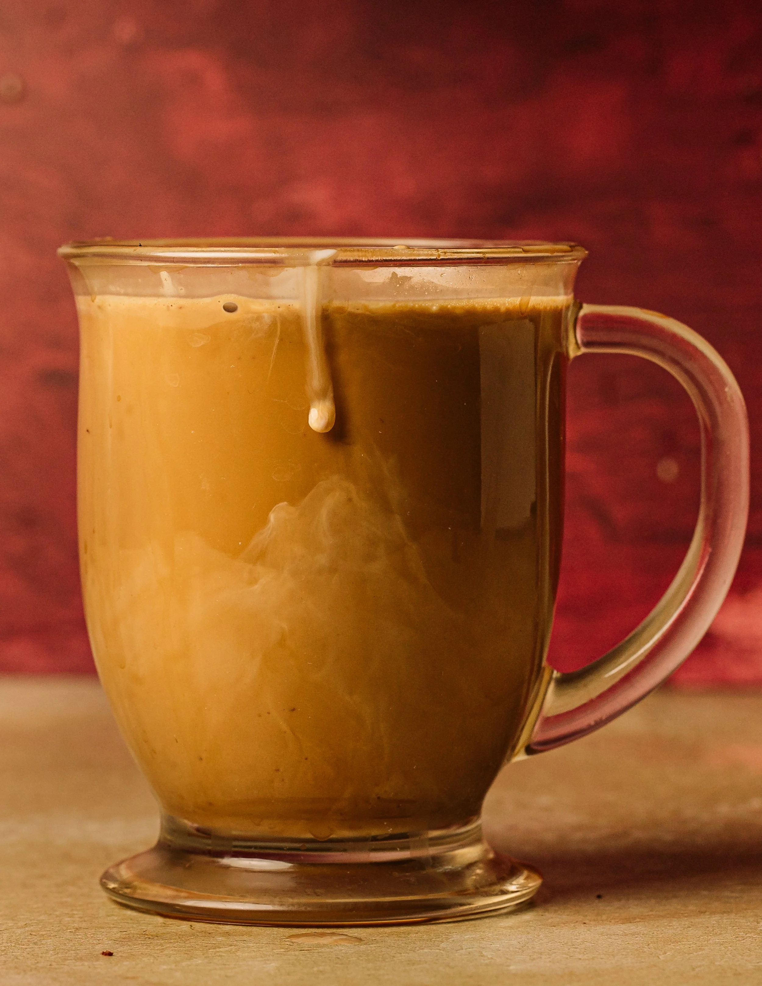 A glass mug filled with a dark hot beverage, with cream or milk being poured into it, creating a swirling pattern.