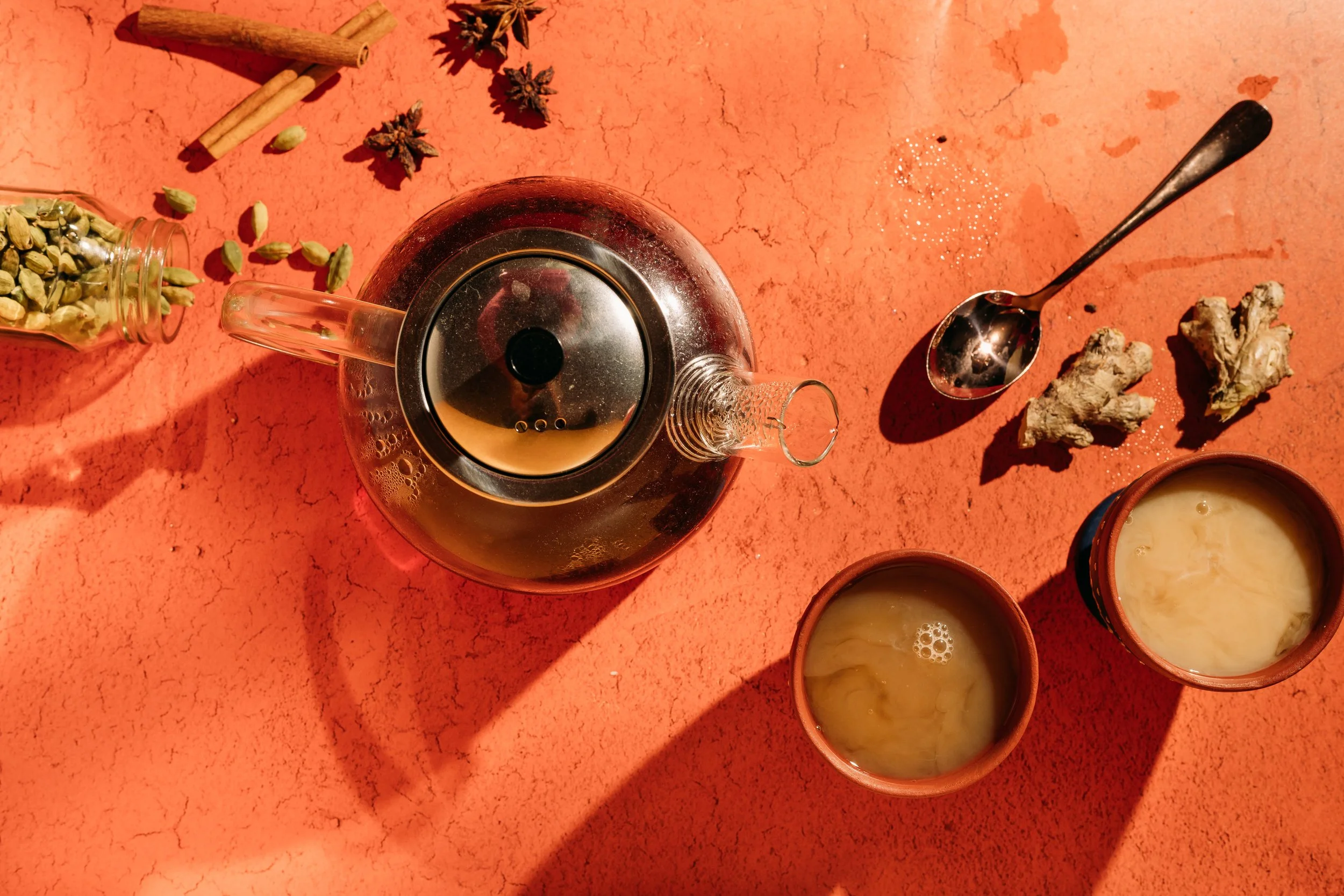 Top-down view of a glass teapot with tea, two small cups of tea, a spoon, ginger roots, cardamom pods, cinnamon sticks, star anise, and scattered peppercorns on a red textured surface.