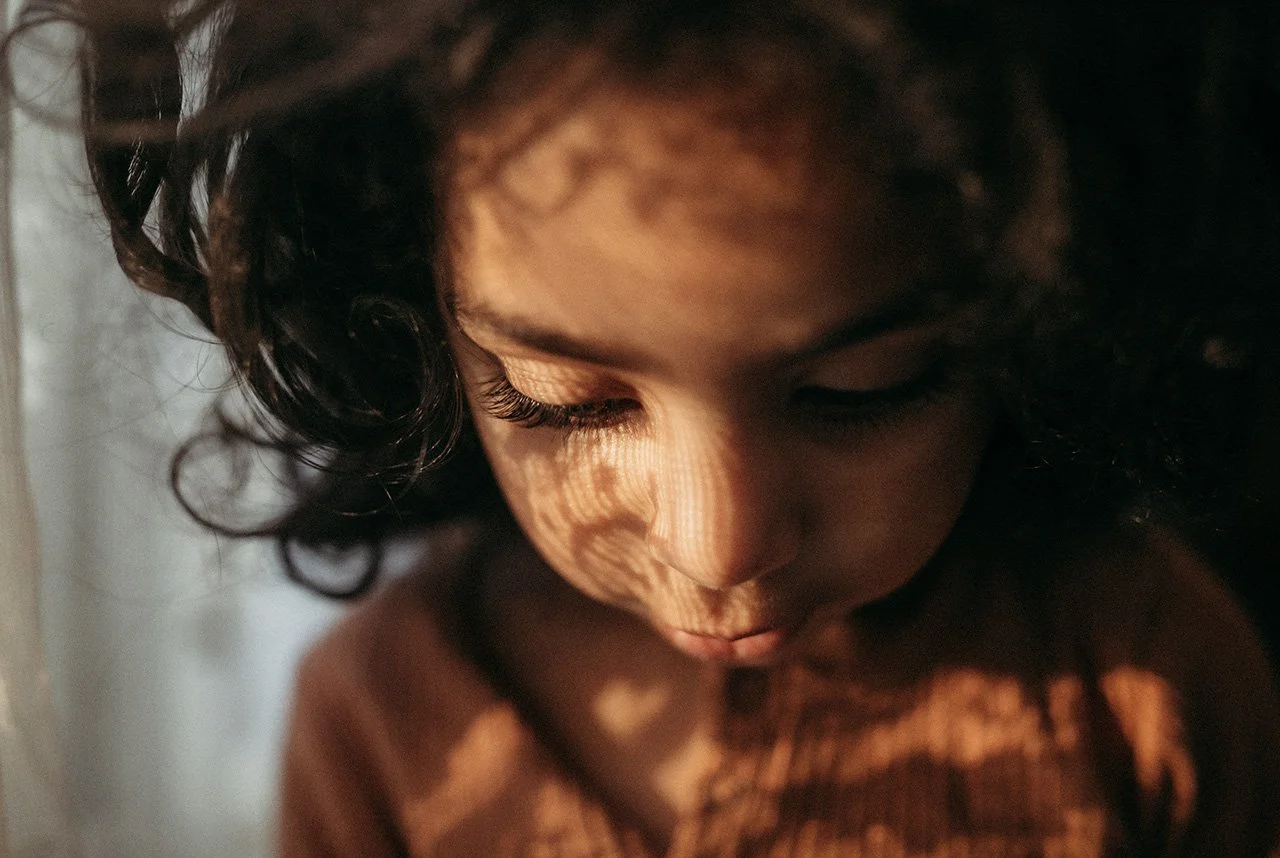 Close-up of a young girl with curly hair and closed eyes, with light casting patterned shadows on her face.