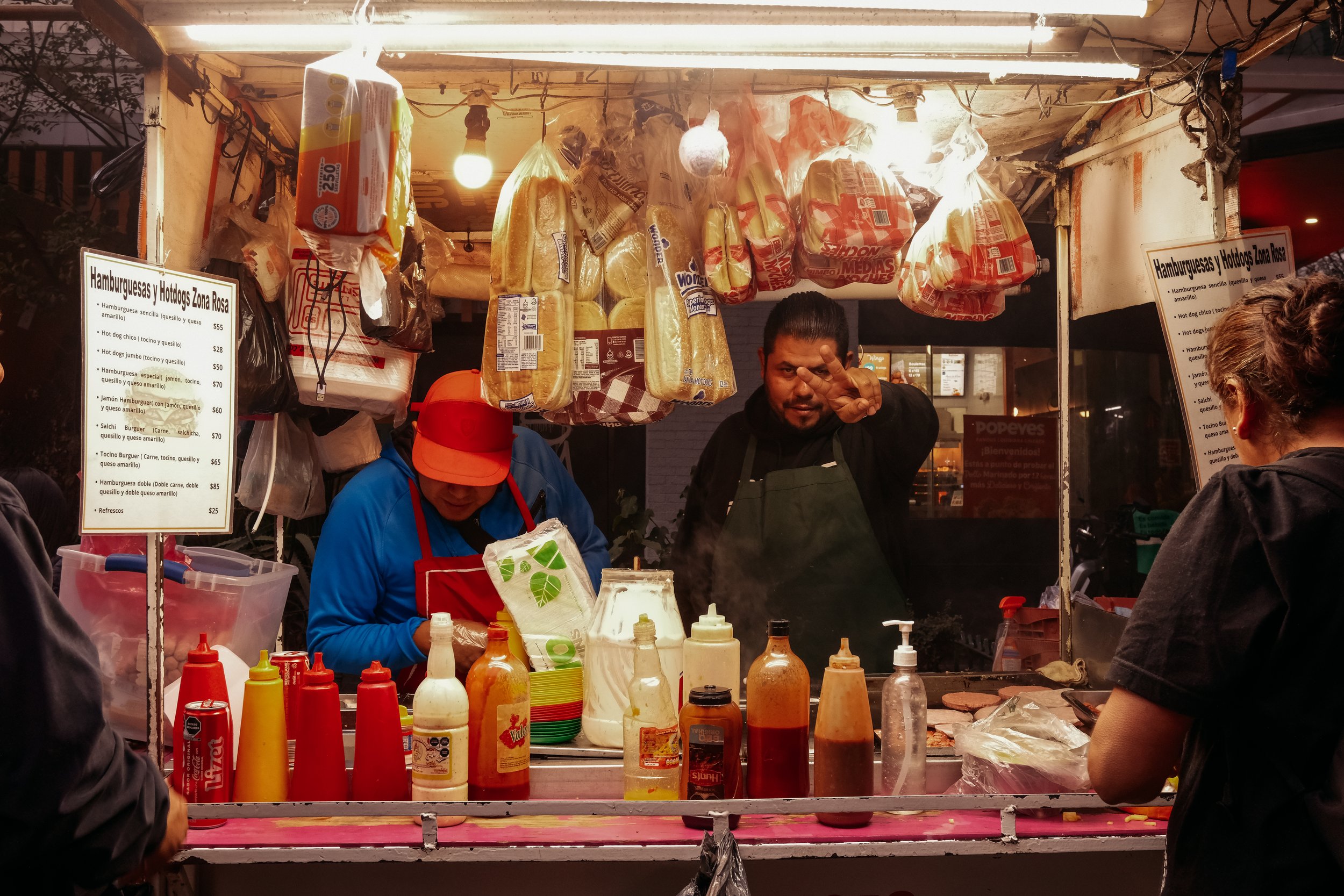 A street vendor stand selling hot dogs, hamburgers, and sandwiches with condiments on a counter. Two vendors are behind the counter, one making a sandwich or hot dog and the other waving at the camera. Customers are in front of the stand ordering.