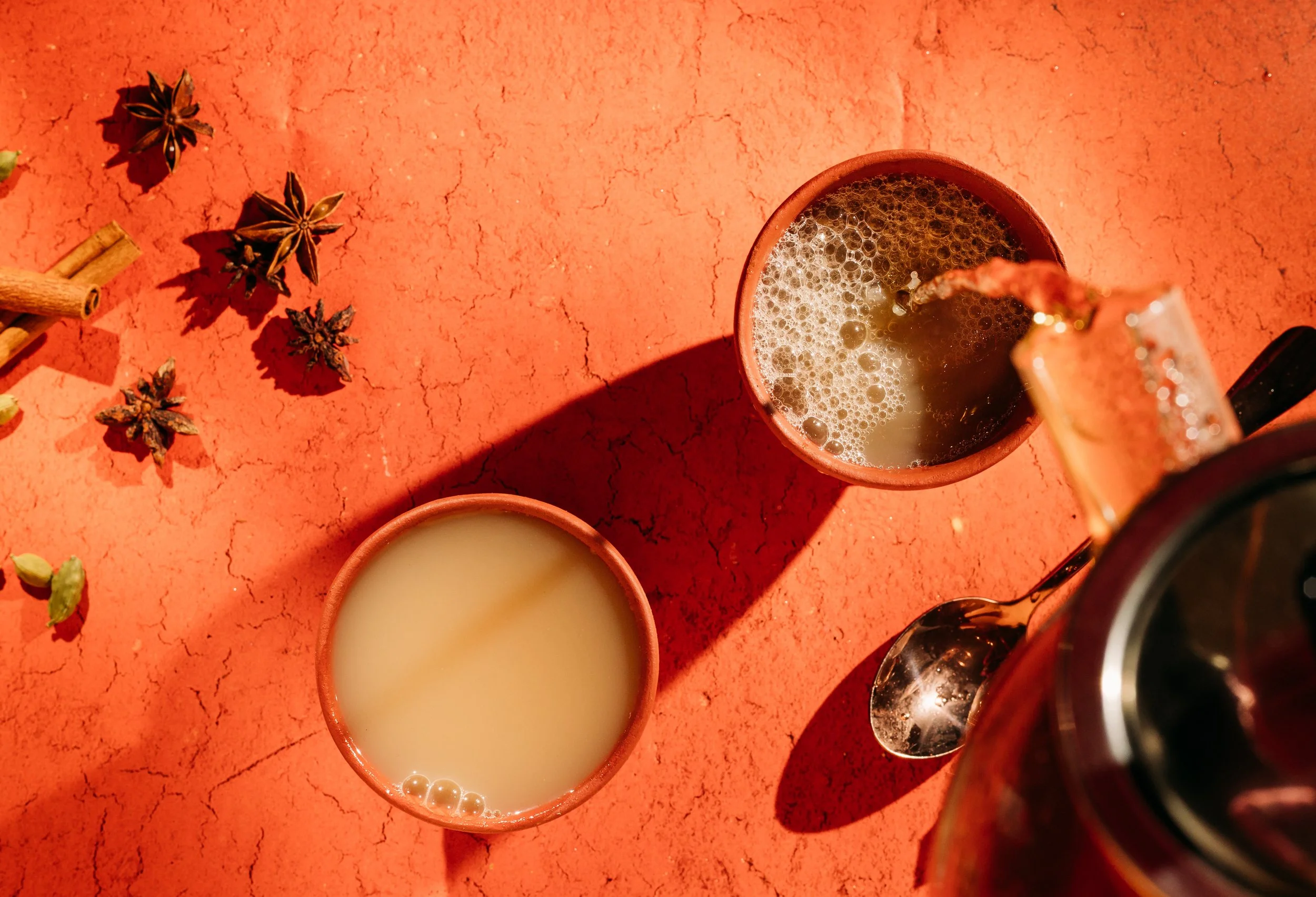 Two cups of tea on a red textured surface, with star anise and cinnamon sticks scattered nearby, and a teapot partially visible.