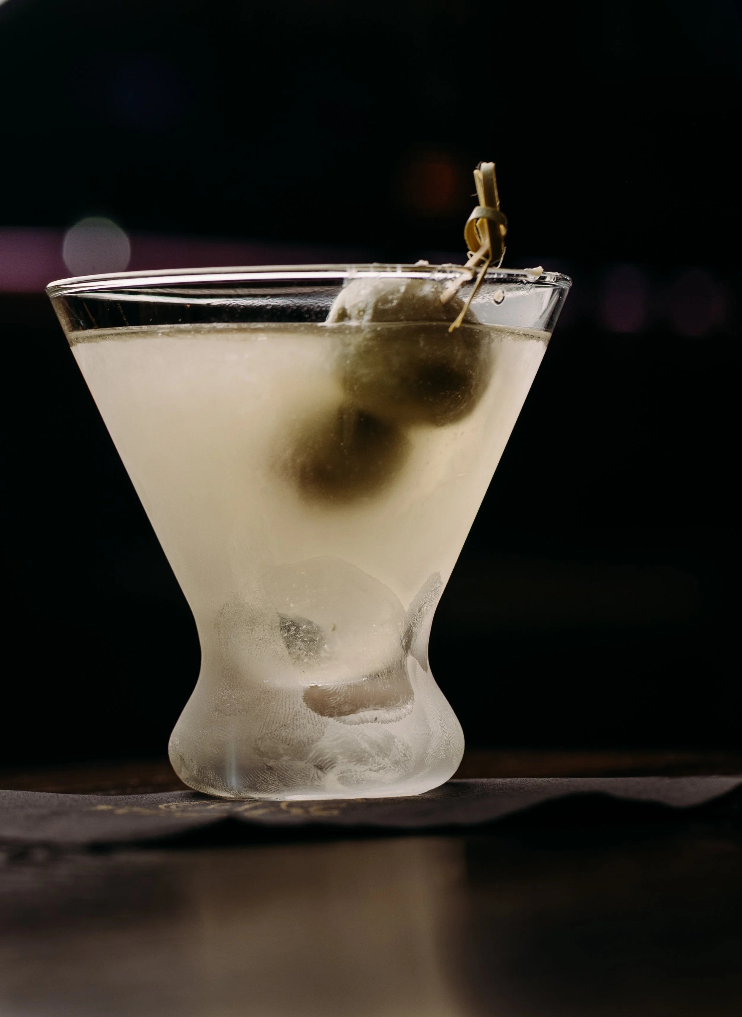 A close-up of a martini glass with a salted rim, filled with a pale yellow cocktail, garnished with green olives on a toothpick, against a dark background.