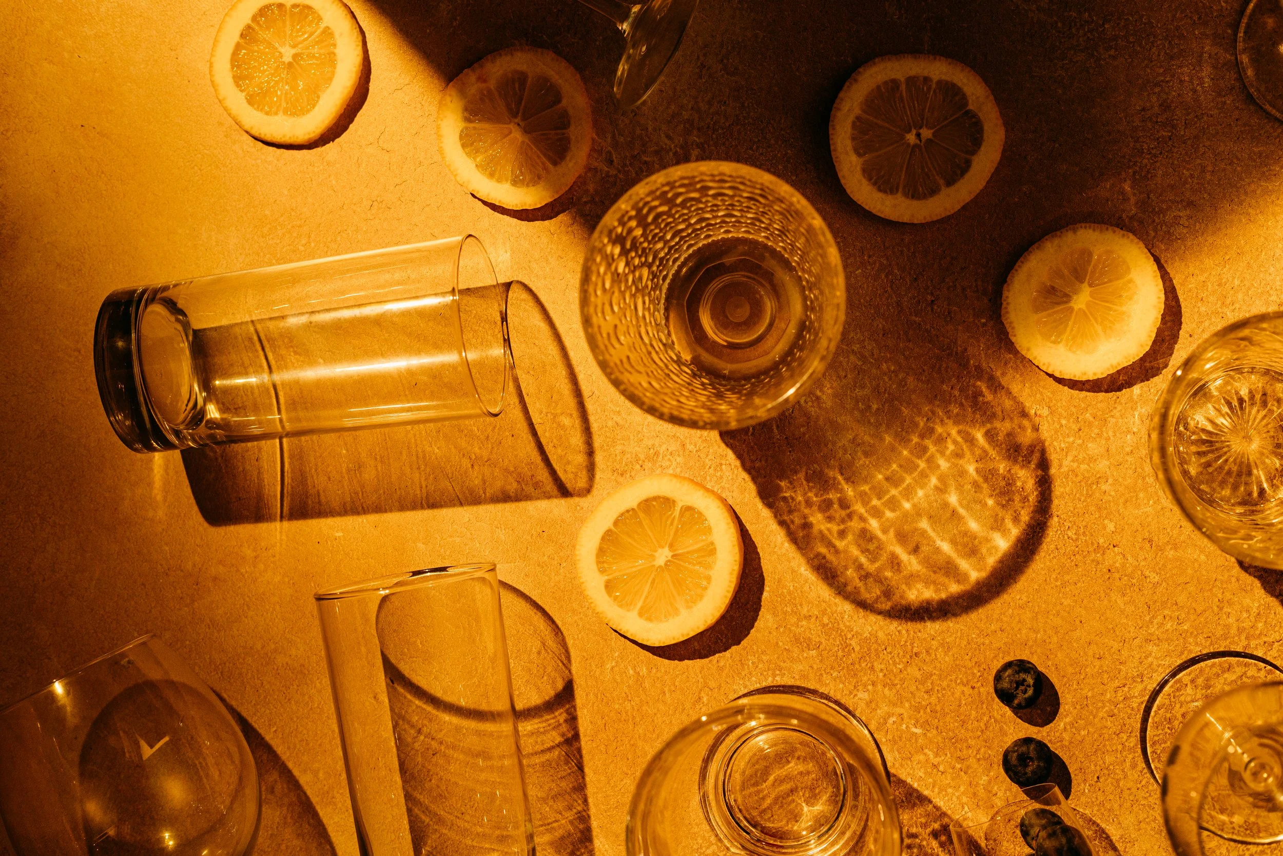 Top-down view of a table with lemon slices, glass cups, and a glass of champagne, illuminated with warm, golden lighting creating shadows and reflections.
